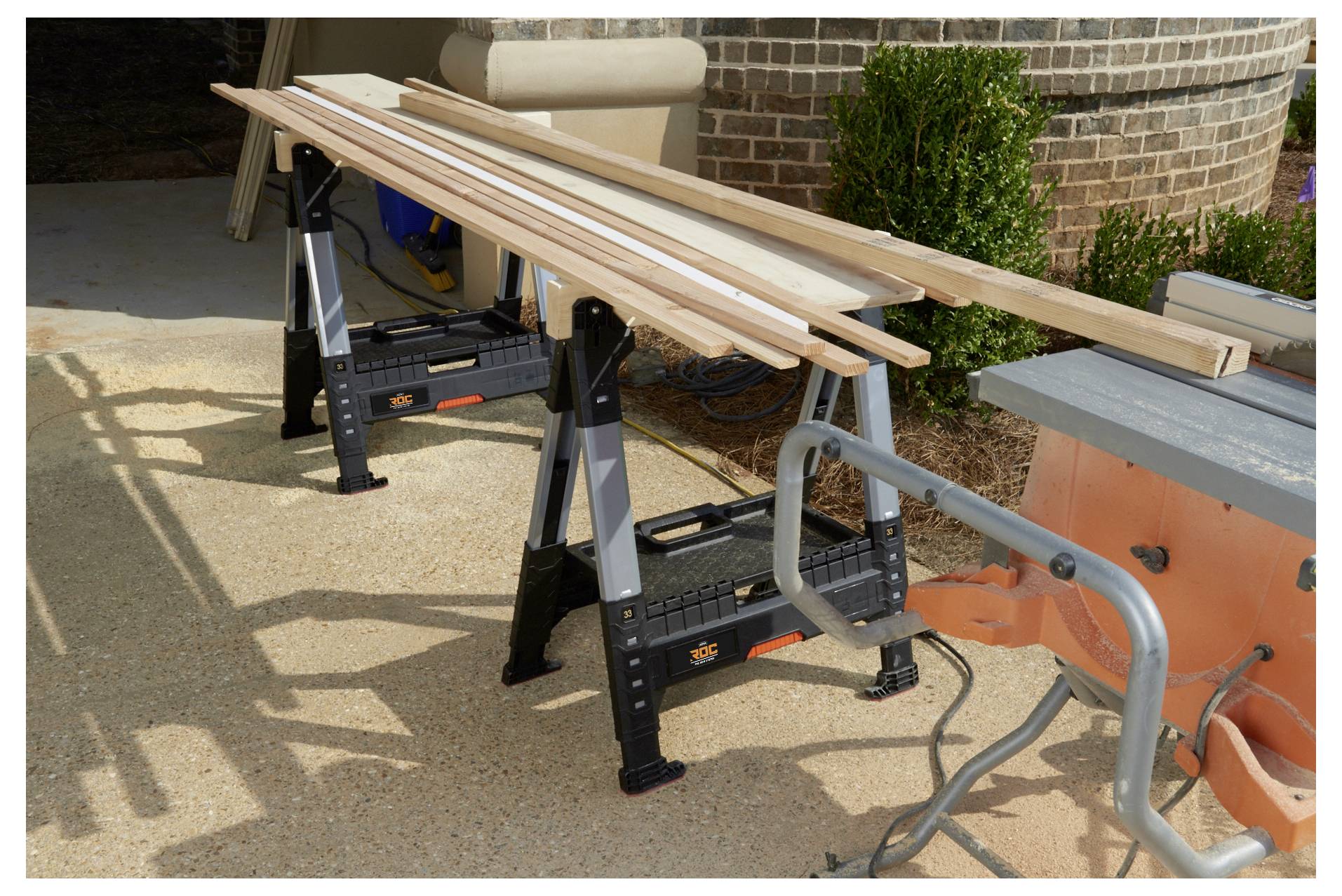 Holzbretter werden auf einem Paar Böcken in einem Außenbereich nahe einer Ziegelwand platziert, wobei eine Tischkreissäge im Vordergrund sichtbar ist.