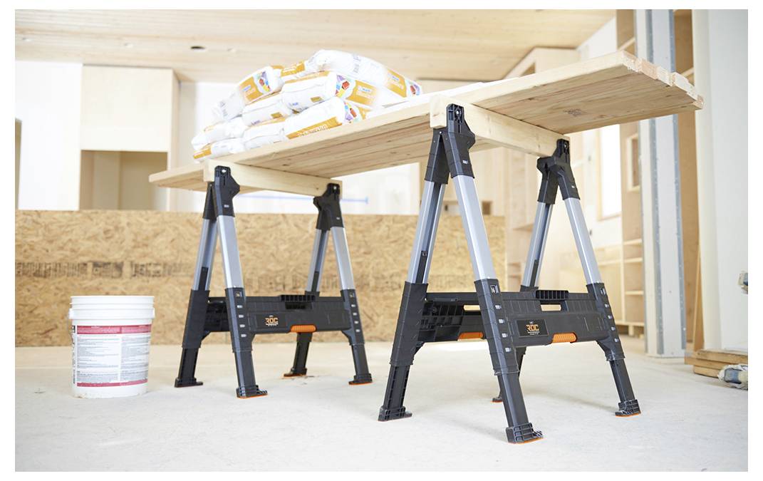 Ein Paar schwarze und orangefarbene klappbare Böcke, die eine Holzplanke abstützen, die mit Säcken von Baumaterialien in einer Baustelle beladen ist.