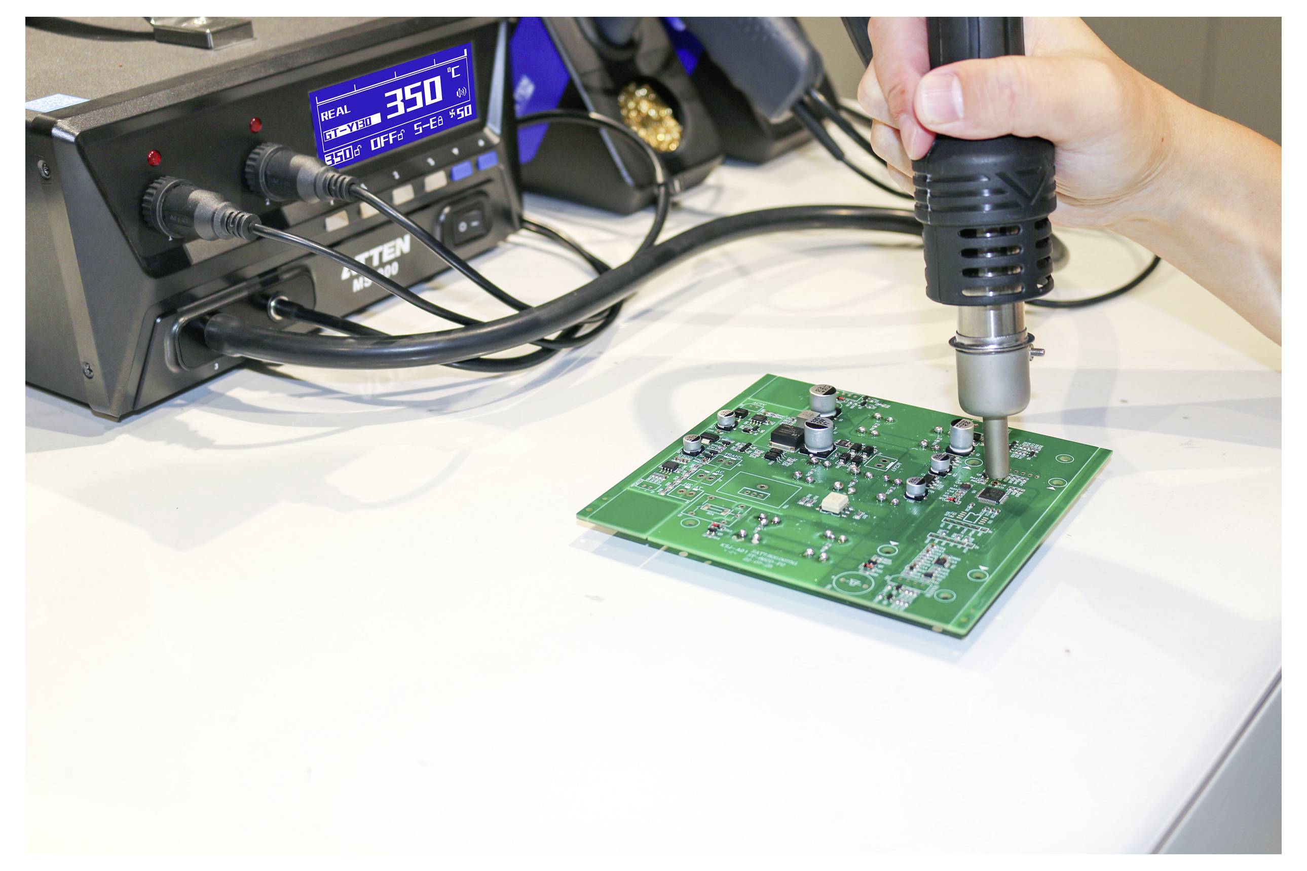Eine Hand, die einen Lötkolben hält, arbeitet an einer grünen Leiterplatte, während im Hintergrund eine digitale Löstation die Temperatur von '350°C' anzeigt.