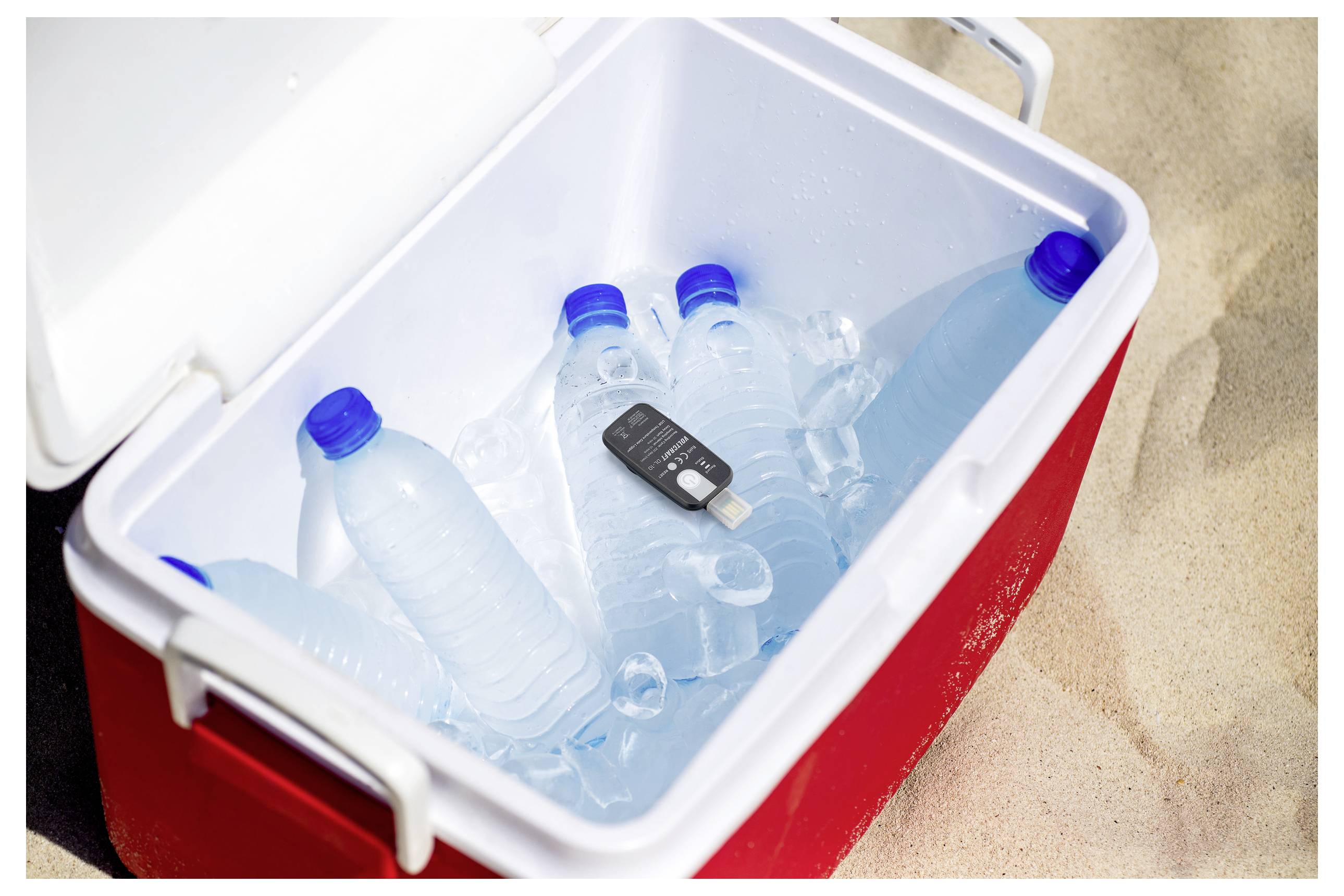 Ein roter Kühlbehälter auf Sand enthält mehrere Kunststoff-Wasserflaschen und einen Autoschlüssel, was ein Strandtags-Setup hervorhebt.