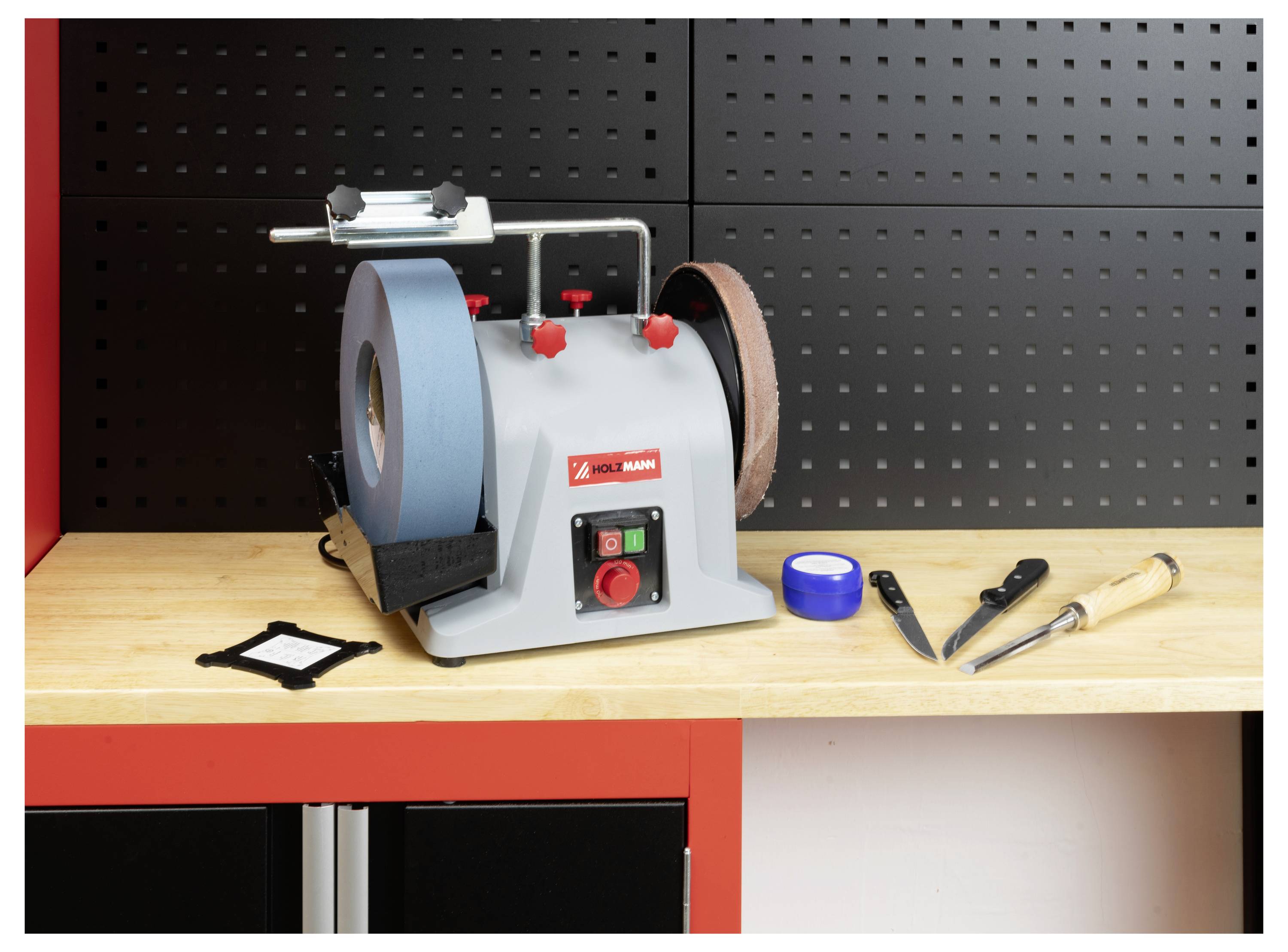 Eine Standschleifmaschine mit einer blauen Schleifscheibe und einer braunen Abziehscheibe steht auf einer Holzwerkbank. In der Nähe liegen ein Meißel, ein Messer und blaues Klebeband.