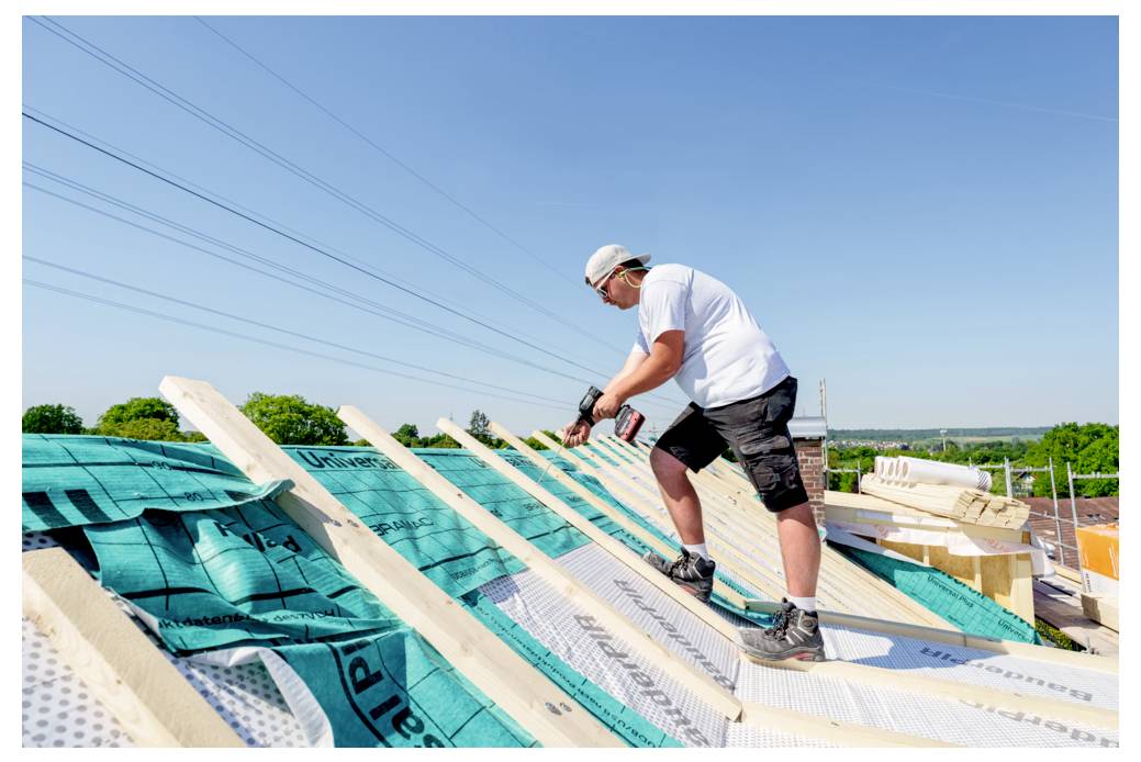 Ein Bauarbeiter in einem weißen Hemd und kurzen Hosen installiert Dachbalken auf einem Dach unter einem klaren blauen Himmel.