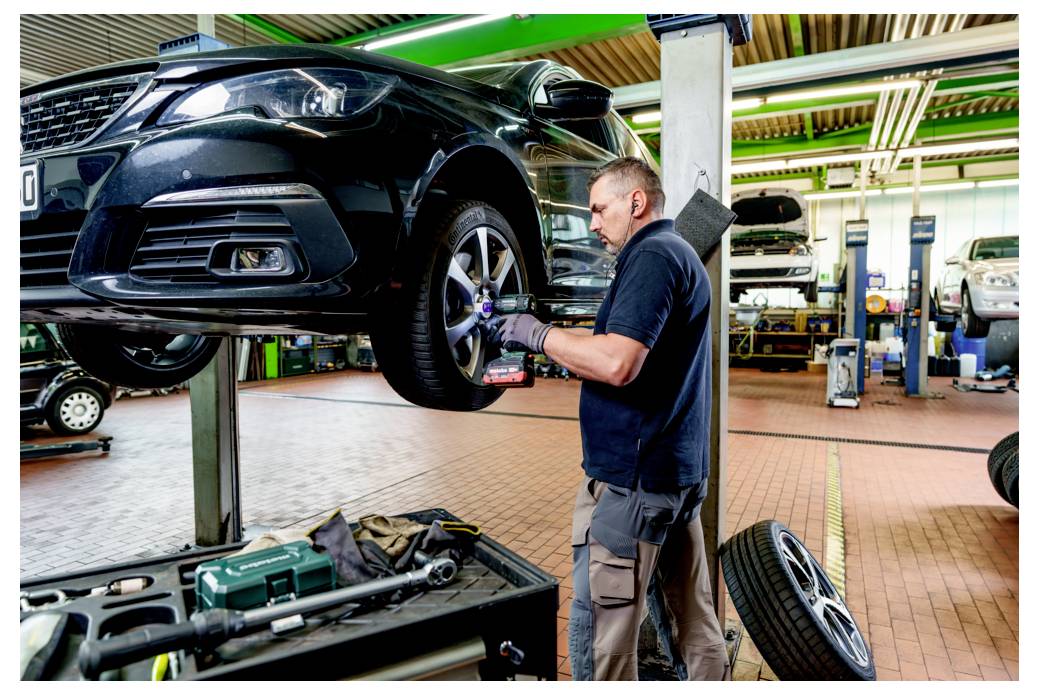 Ein Mechaniker in einer Werkstatt inspiziert das Reifen eines Autos auf einer Hebebühne. Werkzeuge und Ausrüstung sind im Hintergrund der Werkstatt sichtbar.