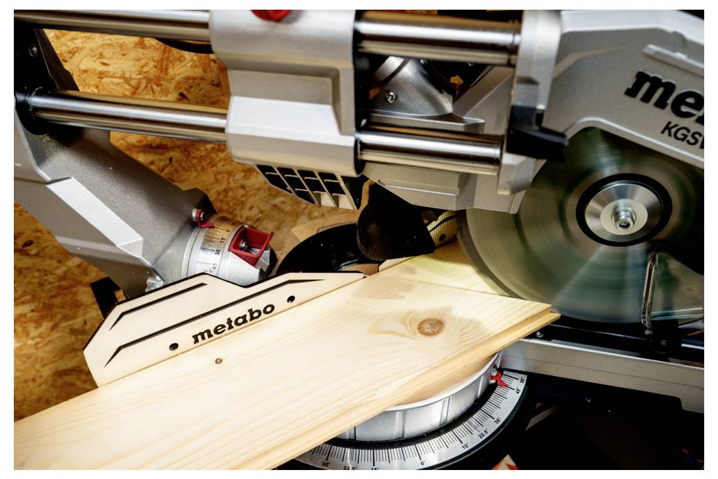 Metabo-Gehrungssäge, die ein Holzbrett in einem Winkel zersägt. Detailaufnahme, die den Kontakt zwischen Sägeblatt und Holz hervorhebt.