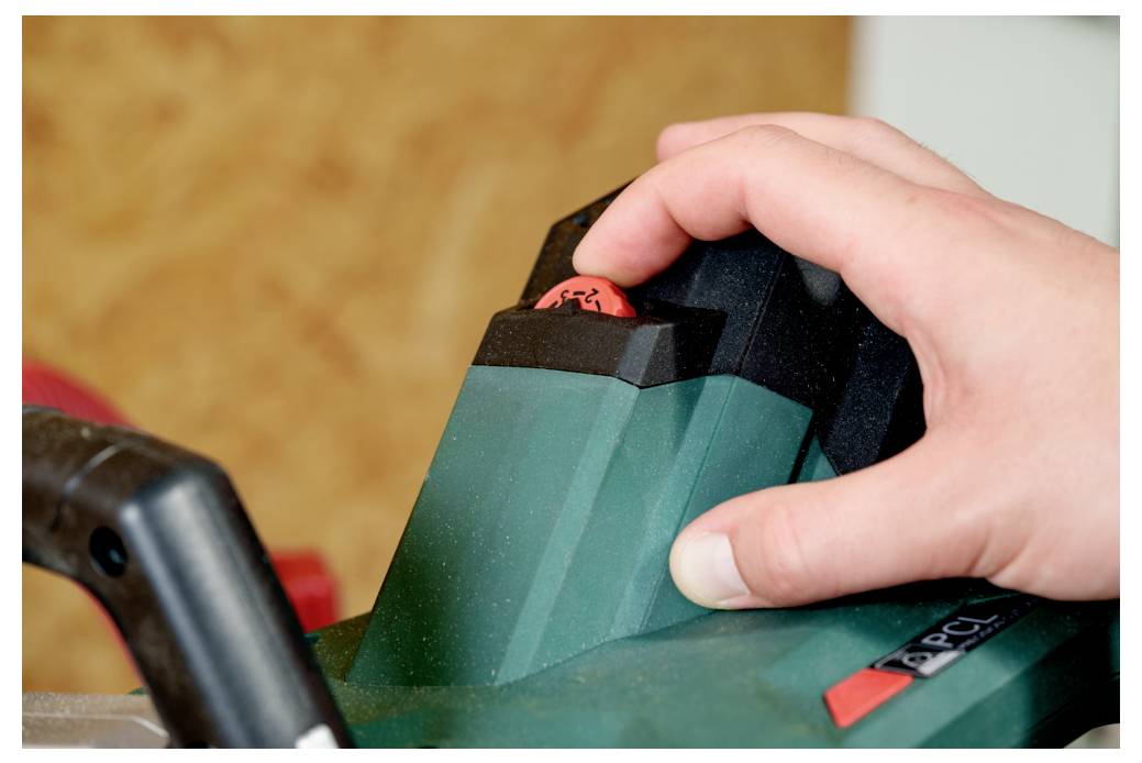 Eine Hand drückt den roten Sicherheitsschalter an einem grünen Elektrowerkzeug und deutet damit auf Aktivierung oder Betrieb in einer Werkstattumgebung hin.