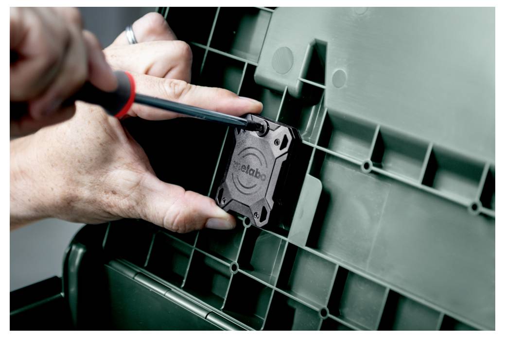 Eine Person verwendet einen Schraubendreher, um eine kleine schwarze Komponente mit der Aufschrift 'Metabo' auf eine grüne Kunststoffoberfläche mit Gittermuster zu befestigen.