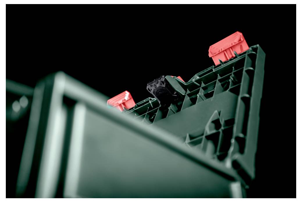 Eine Plastikkiste, teilweise geöffnet, die ein strukturiertes Innenleben und drei rote Verschlüsse vor einem schwarzen Hintergrund zeigt.