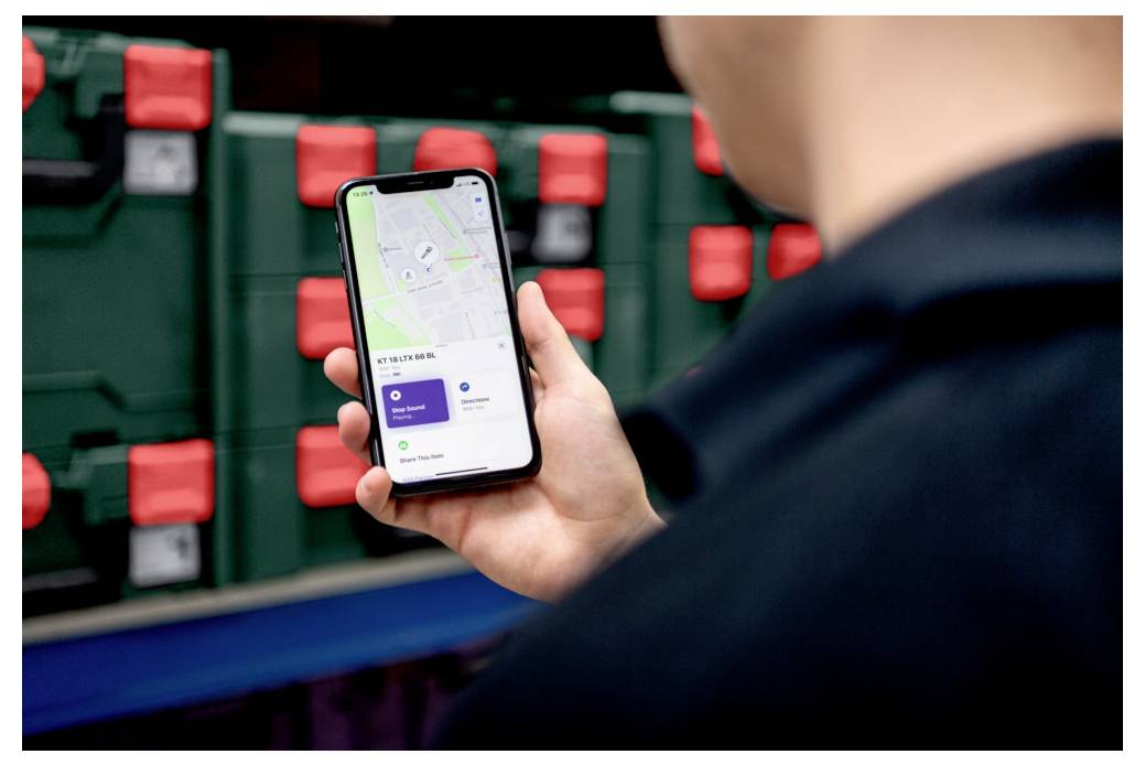 Eine Person hält ein Smartphone, auf dem eine Karte und Standortverfolgungsinformationen angezeigt werden. Im Hintergrund befinden sich Regale mit grünen und roten Kisten.