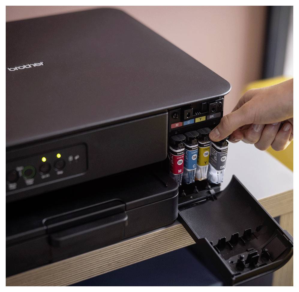 Eine Hand setzt eine gelbe Tintenpatrone in den Tinteneinschub eines Brother-Druckers, neben den Patronen in Cyan, Magenta und Schwarz.