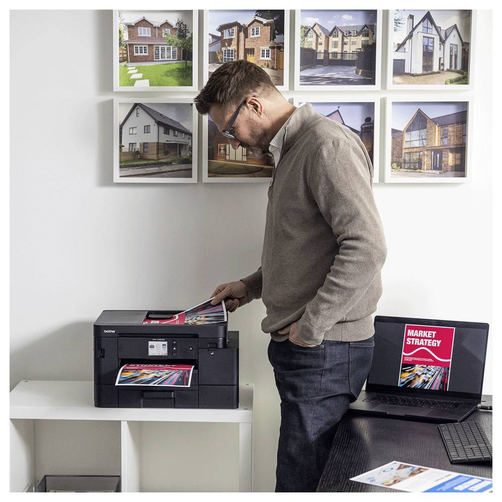 Ein Mann bedient einen Drucker in einem Büro, umgeben von gerahmten Fotos von Häusern. Ein Laptop zeigt ein Dokument mit dem Titel 