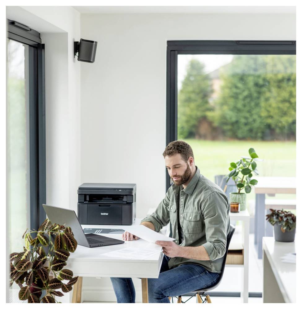 Ein Mann sitzt an einem Schreibtisch in einem hellen Büro, arbeitet an einem Laptop und schaut Dokumente durch, umgeben von Pflanzen und einem Drucker.