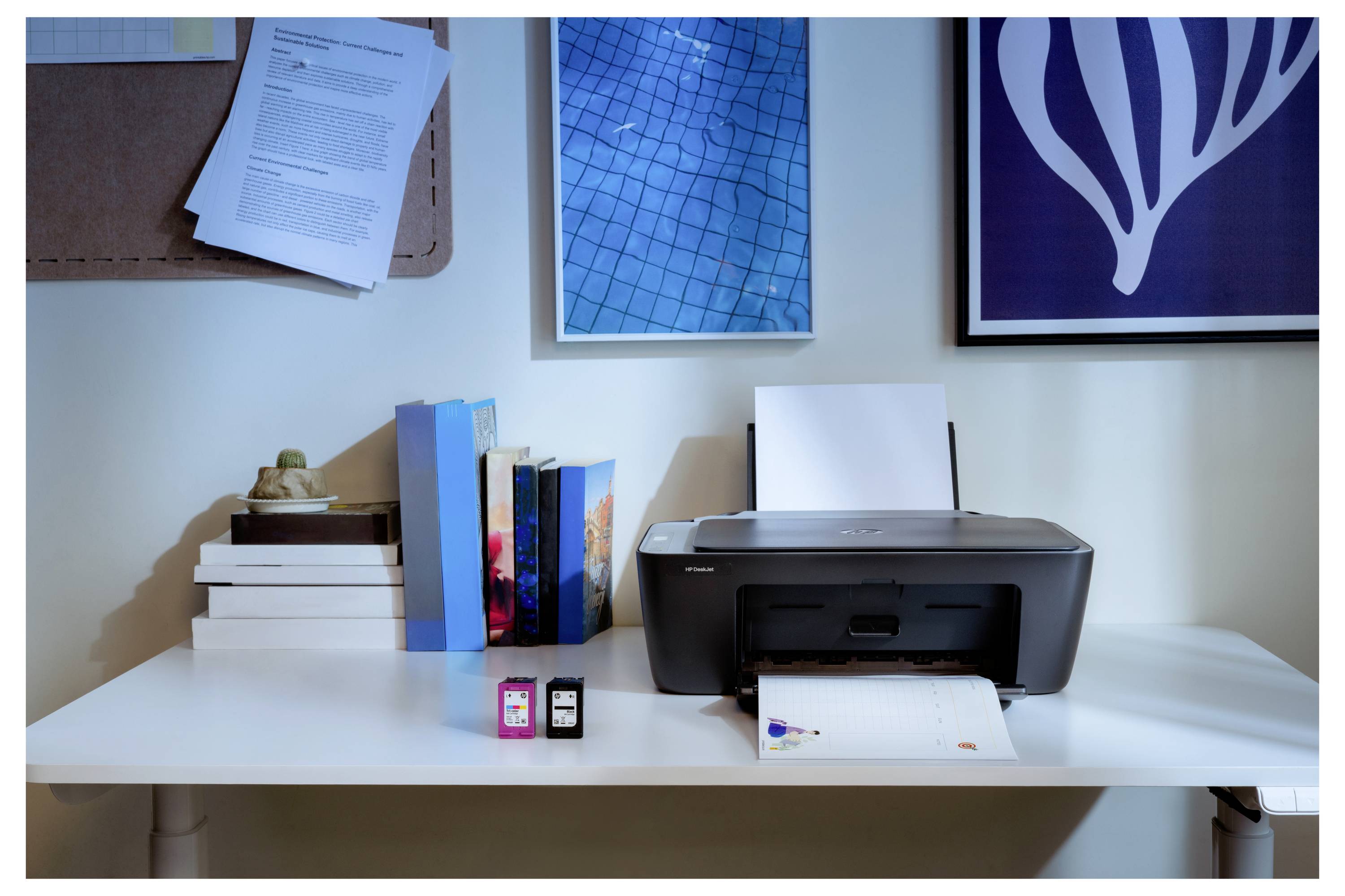 Ein Home-Office-Schreibtisch mit einem Drucker, Tintenpatronen, gestapelten Büchern und dekorativer Wandkunst im Hintergrund.