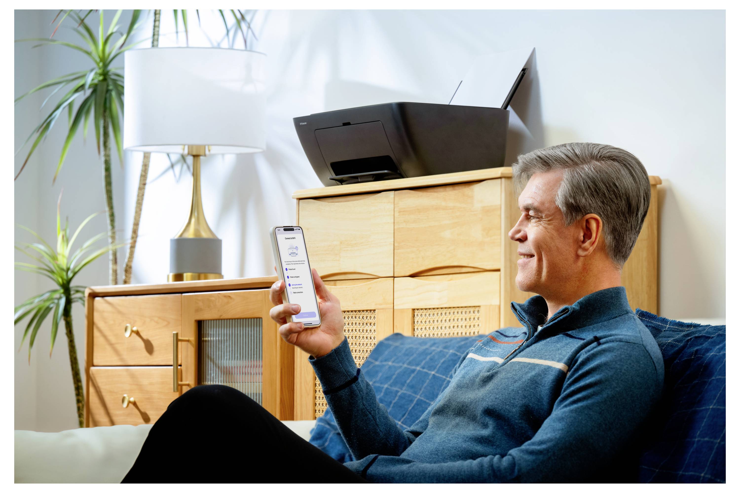 Ein Mann sitzt auf einem Sofa und lächelt, während er auf ein Smartphone schaut. Ein Multifunktionsdrucker ist im Hintergrund sichtbar. Die Umgebung ist ein gemütliches Wohnzimmer.