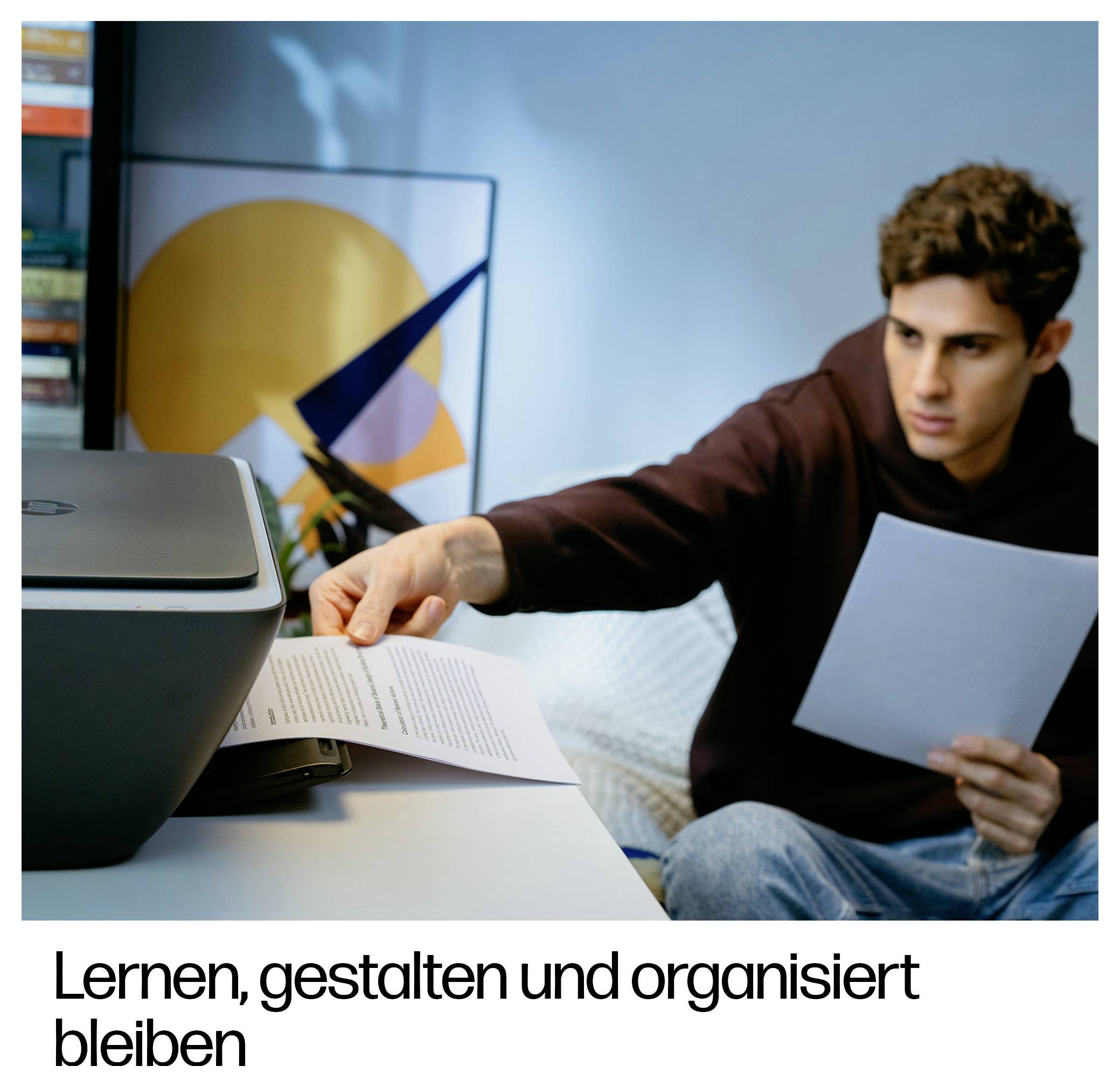 Ein junger Mann sitzt an einem Schreibtisch, hält ein Blatt Papier und füttert einen Drucker. Im Hintergrund ein abstraktes Kunstwerk.