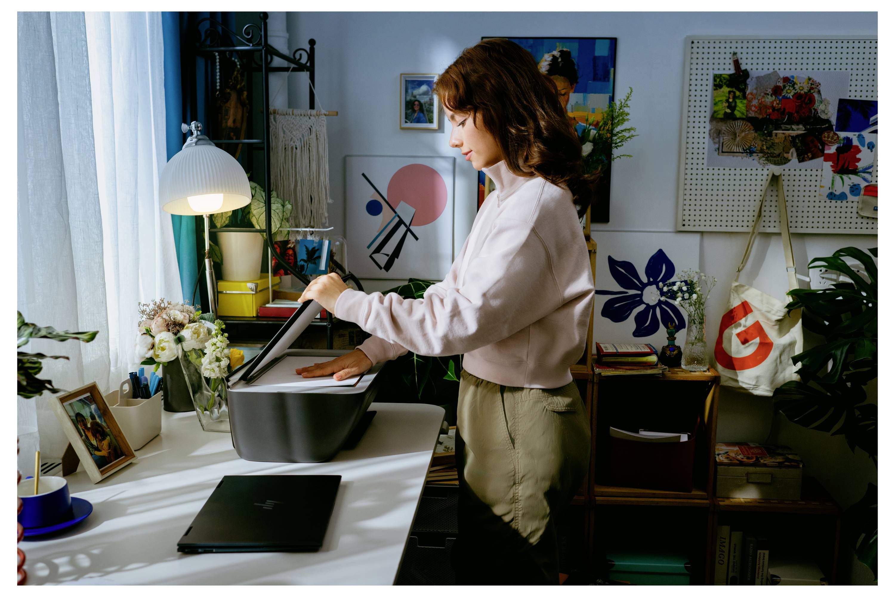 Frau, die einen Tischscanner auf einem Schreibtisch in einem Heimbüro öffnet, mit Laptop, Lampe, Blumen und Kunstwerken in der Nähe.