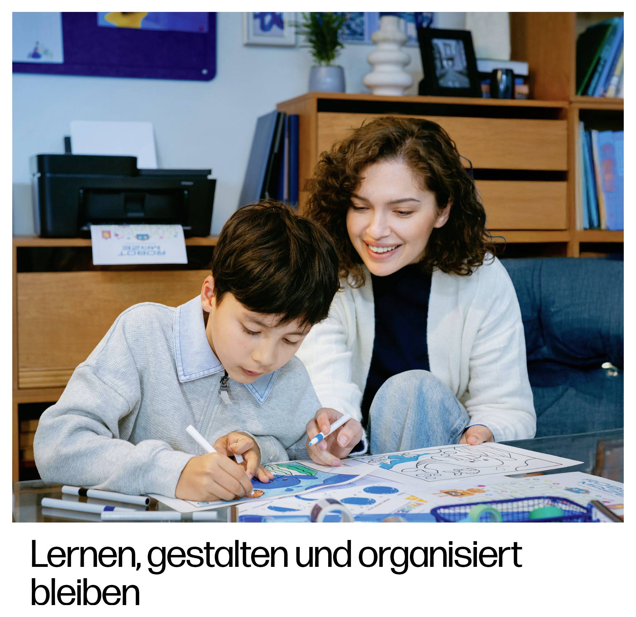 # Lernen, gestalten und organisiert bleibenEine Frau hilft einem Jungen, auf Papieren zu malen und zu zeichnen; der deutsche Text darunter lautet „Lernen, gestalten und organisiert bleiben"