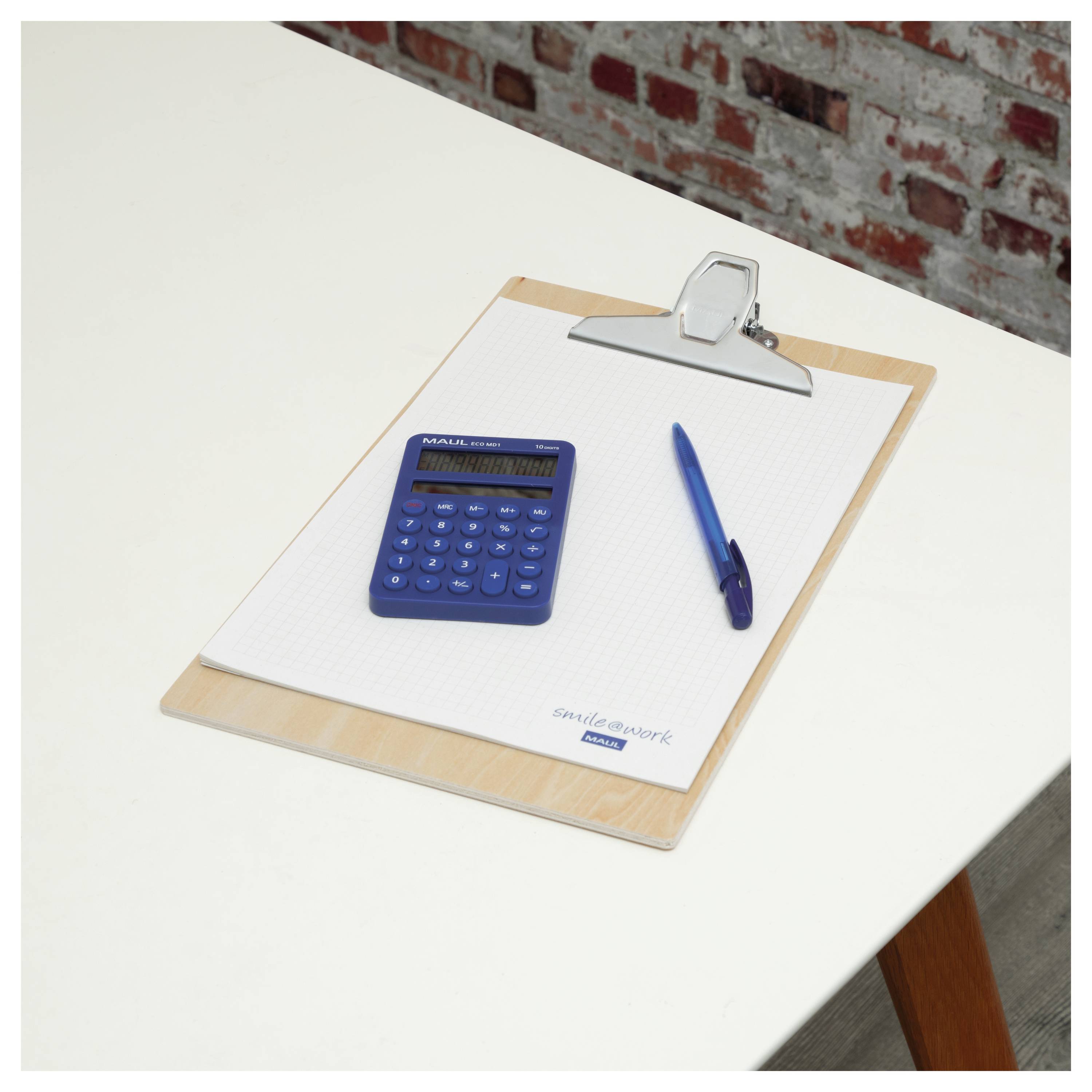 Eine Zwischenablage auf einem weißen Tisch hält einen Notizblock mit einem blauen Taschenrechner und einem Stift darauf, vor einem Ziegelwandhintergrund.