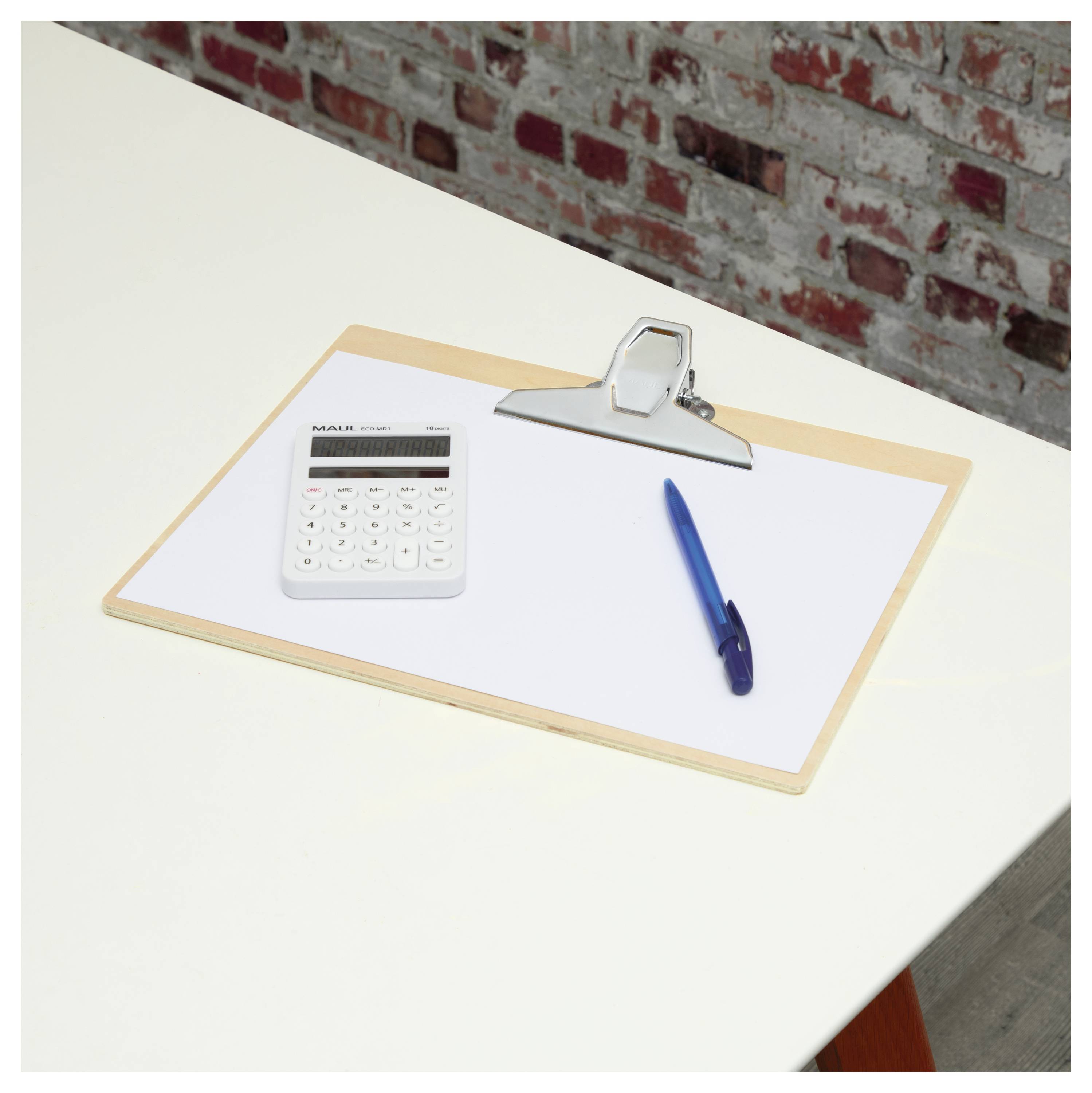 Eine Zwischenablage hält ein leeres Blatt Papier auf einem weißen Schreibtisch, mit einem blauen Stift und einem weißen Taschenrechner daneben, vor einer Ziegelwand im Hintergrund.