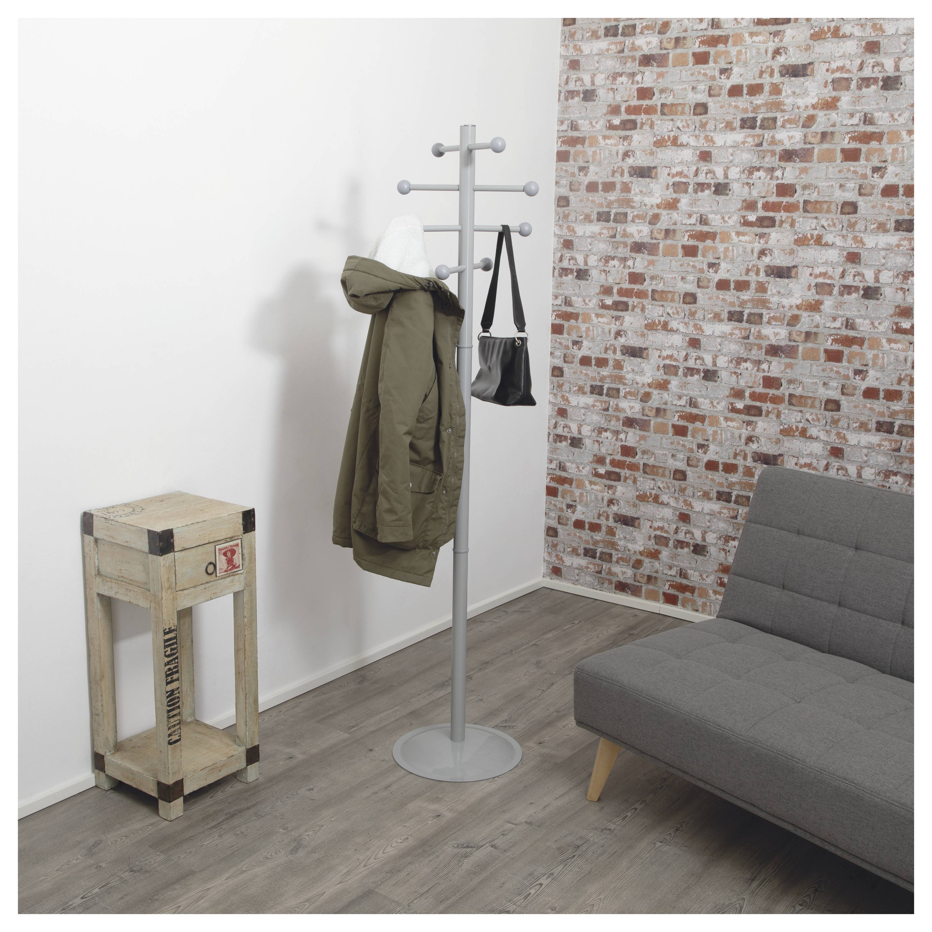 Flur mit einer Garderobe, an der eine grüne Jacke und eine schwarze Tasche hängen, einem kleinen Holzständer und einem grauen Sofa vor einer Mauer aus Backsteinen und weißer Wand.