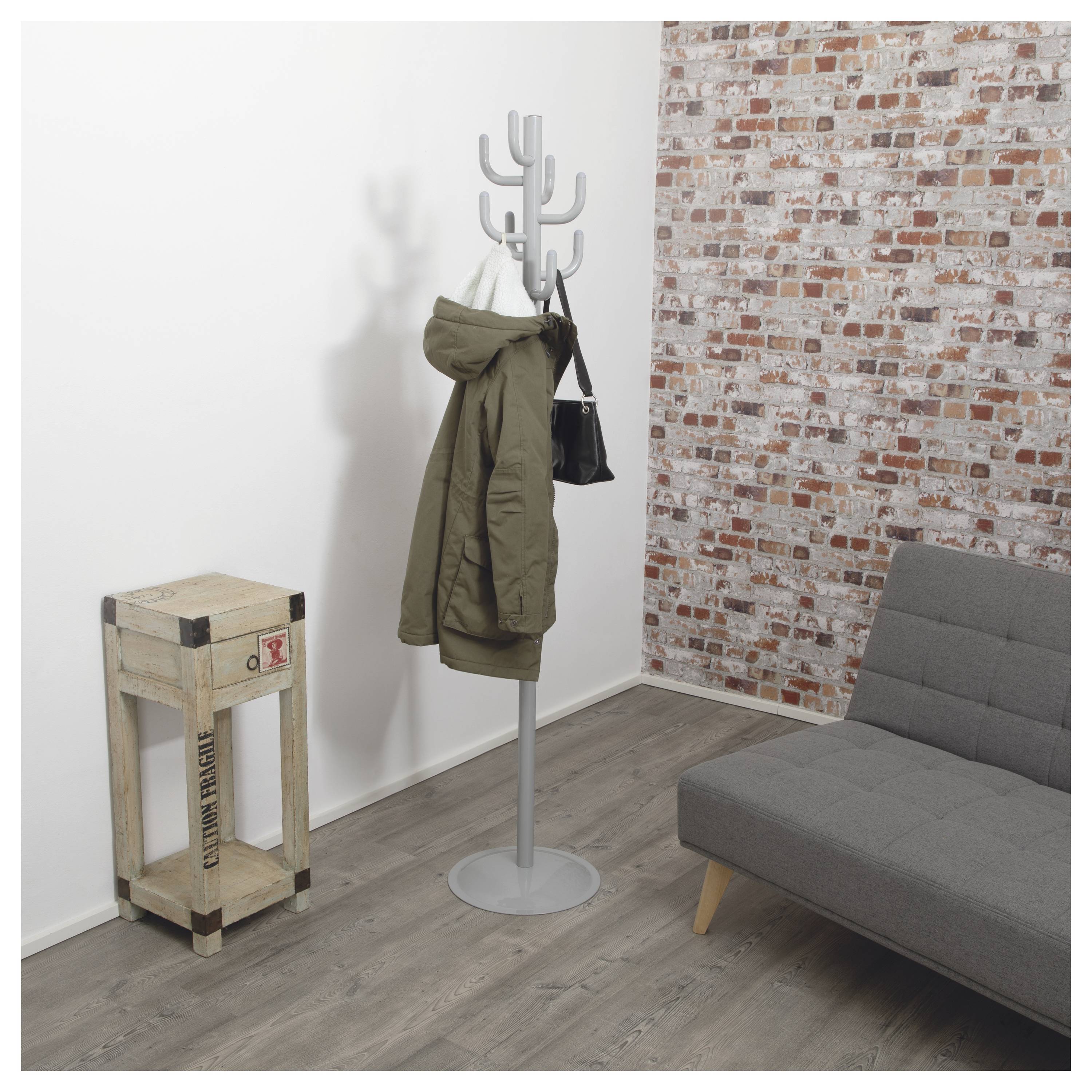 Ein Raum mit einer grauen Garderobe, an der eine olivgrüne Jacke und eine kleine schwarze Tasche hängen, eine Ziegelwand, ein Holzhocker und ein grauer Sofa.