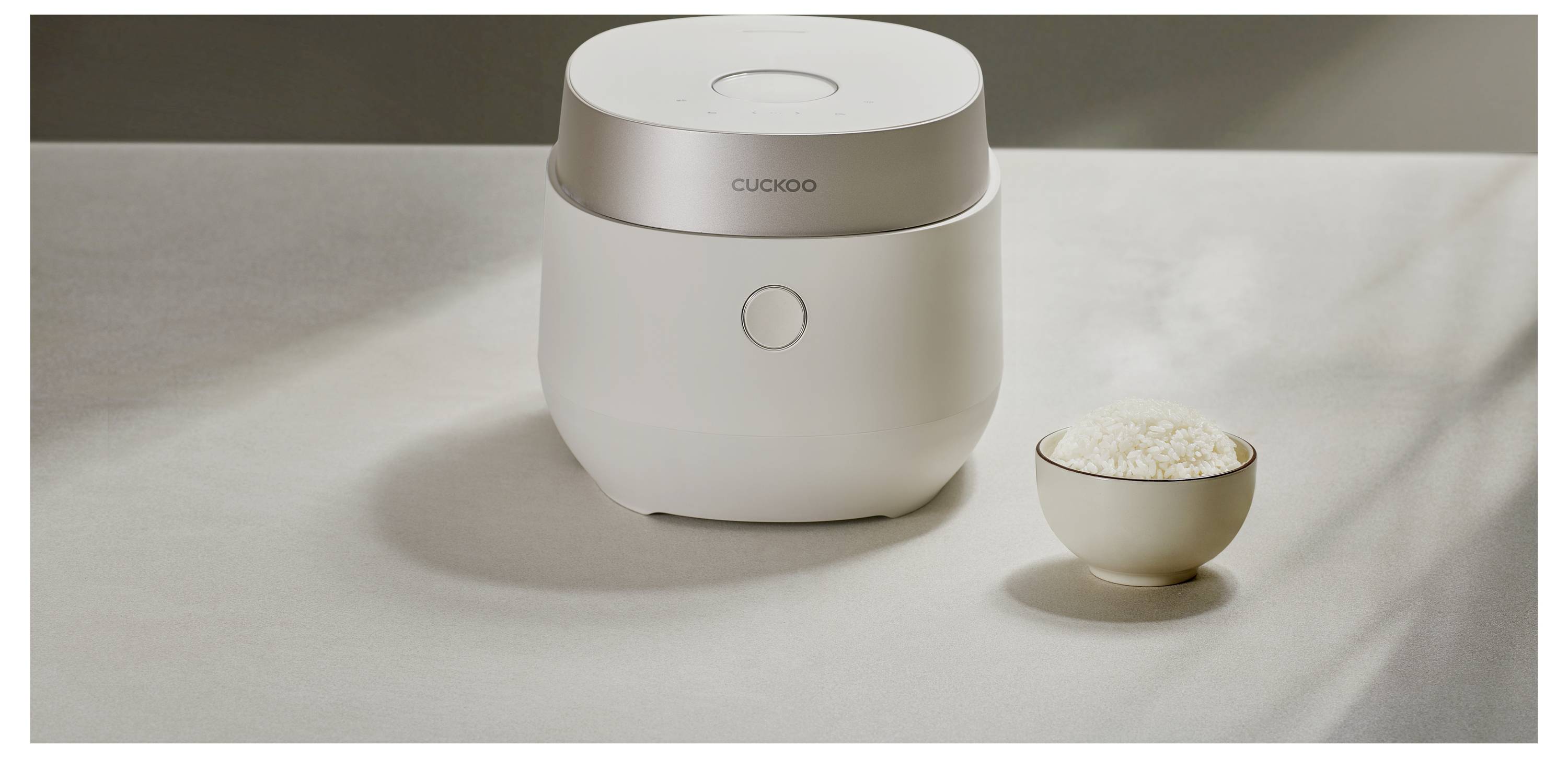 Ein schlanker weißer Reiskocher auf einem Tisch neben einer Schüssel gefüllter gedämpfter Reis.