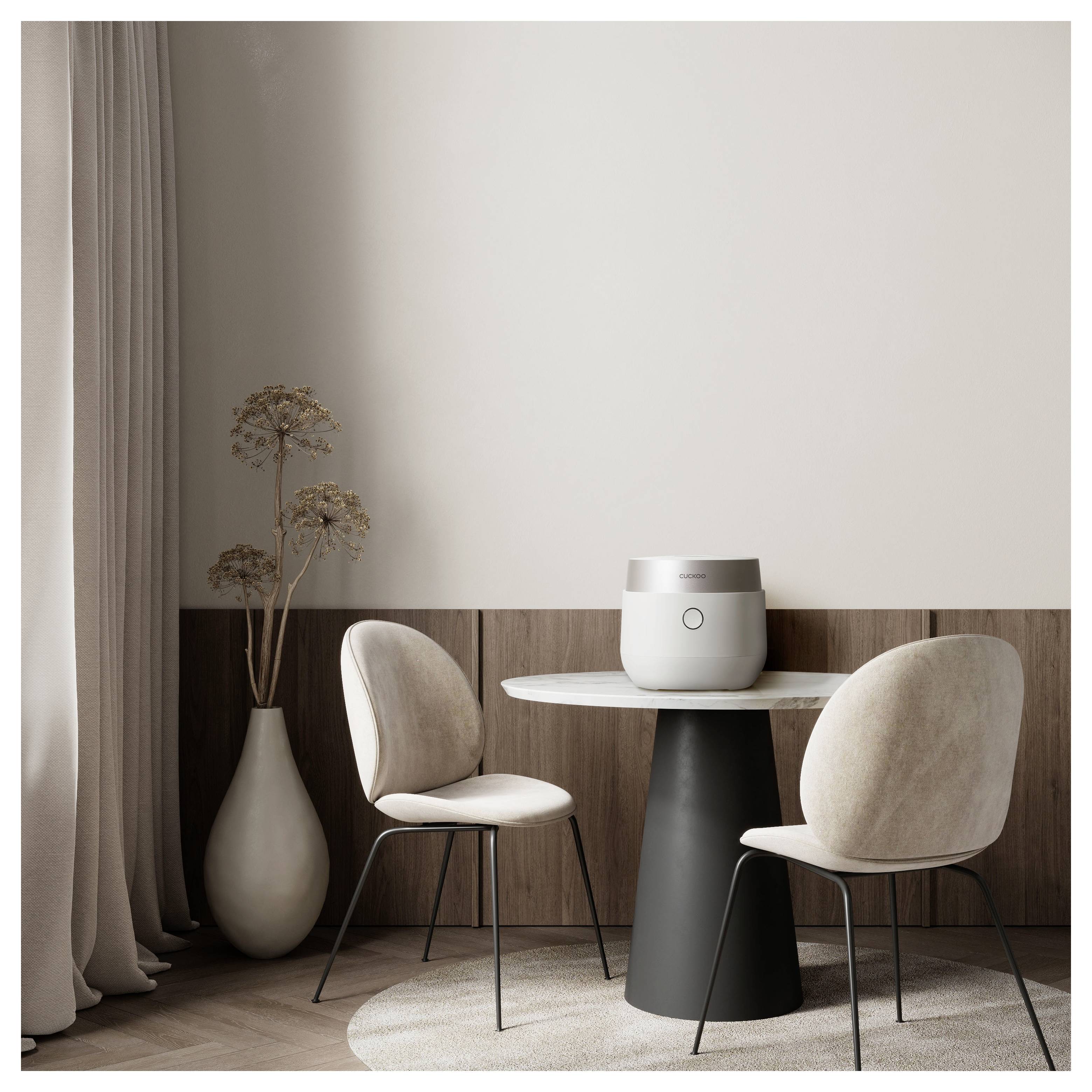 Ein minimalistischer Essbereich mit einem kleinen runden Tisch, zwei beigen Stühlen, einer Topfpflanze und einem runden Gerät auf dem Tisch vor einer neutralen Wand.