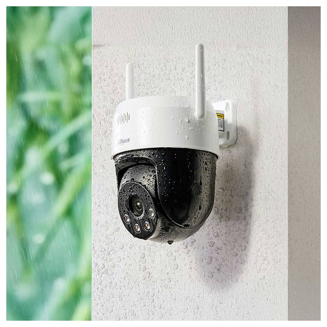 Eine weiße und schwarze Outdoor-Sicherheitskamera, die an einer Wand befestigt ist, mit Wassertropfen bedeckt und mit einem unscharfen grünen Hintergrund links davon.