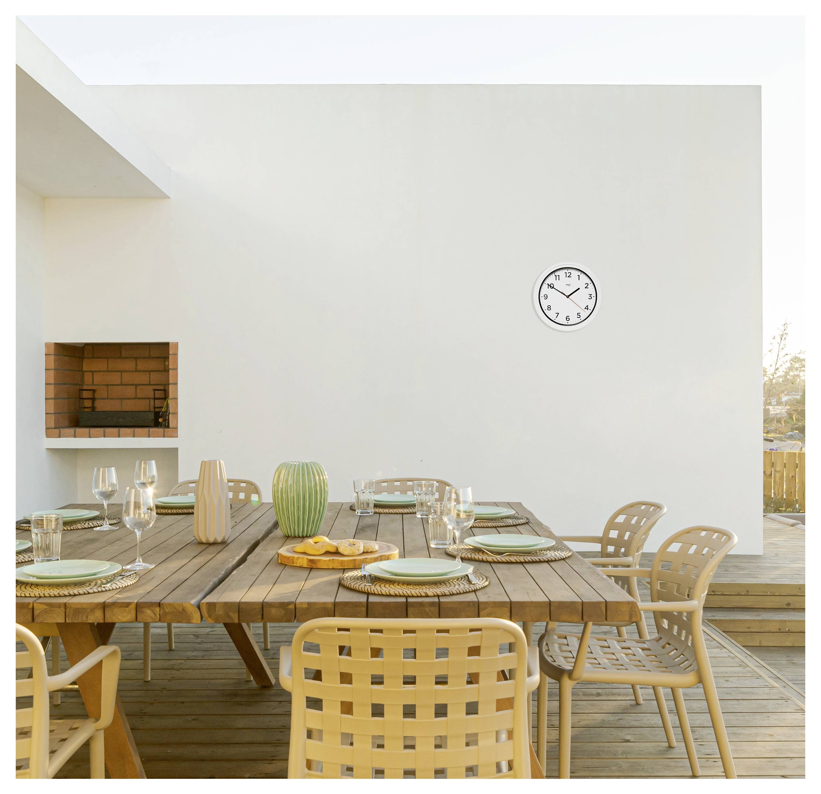 Eine moderne Outdoor-Essbereich mit einem Holztisch, der für sechs Personen gedeckt ist, mit Tellern und Gläsern. Eine Uhr zeigt 7:00 Uhr an einer weißen Wand. Ein Grill ist eingebaut.