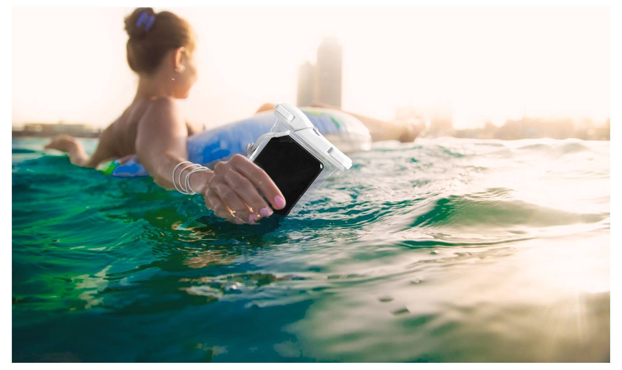 Eine Person, die auf dem Wasser treibt, hält ein wasserfestes Klapphandy dicht an die Wasseroberfläche. Die Sonne geht unter und wirft einen warmen Glanz über die Skyline der Stadt im Hintergrund.