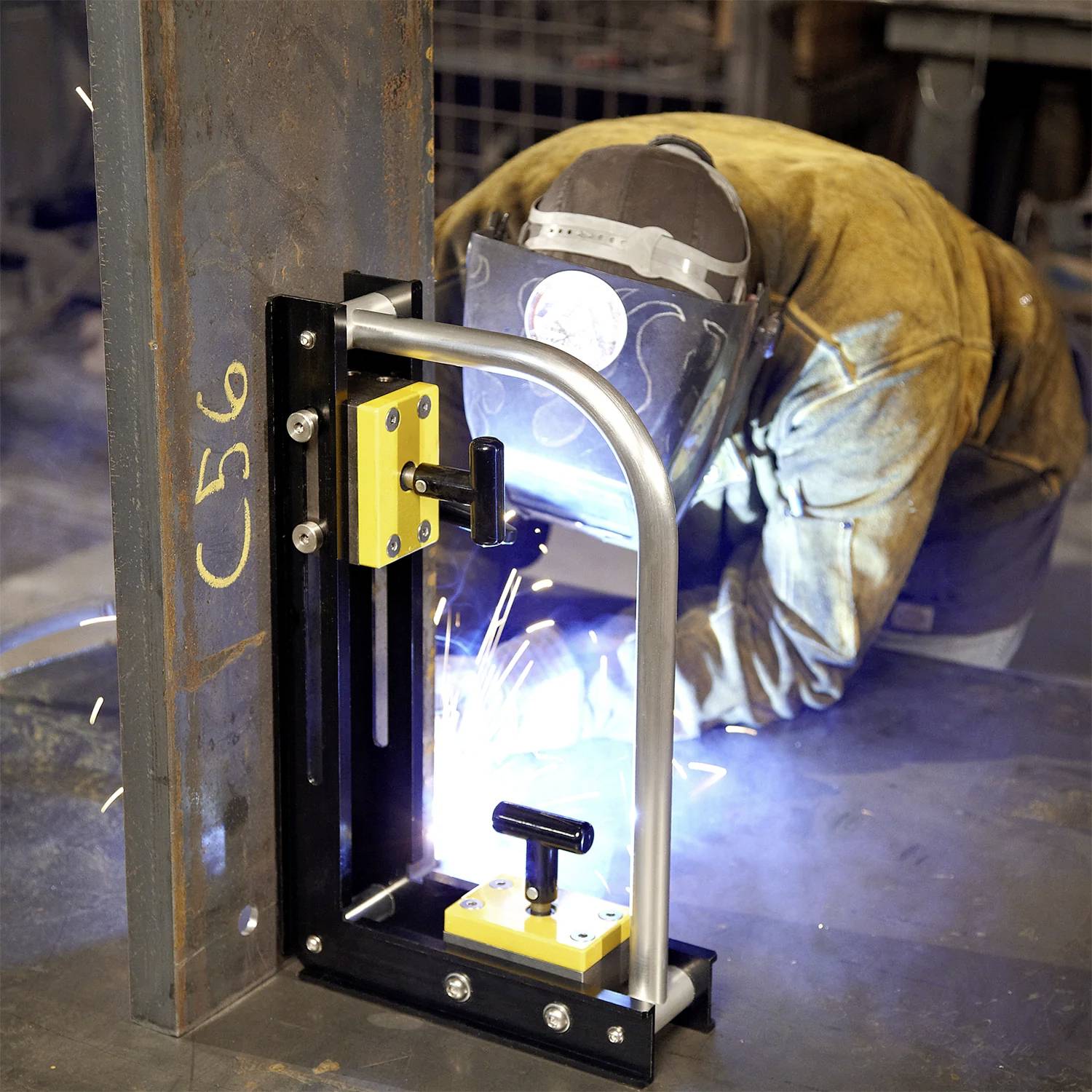 Eine Person mit Schutzausrüstung schweißt Metall in einem industriellen Umfeld, wobei Funken und ein Stahlrahmen mit der Markierung 'C56' sichtbar sind.