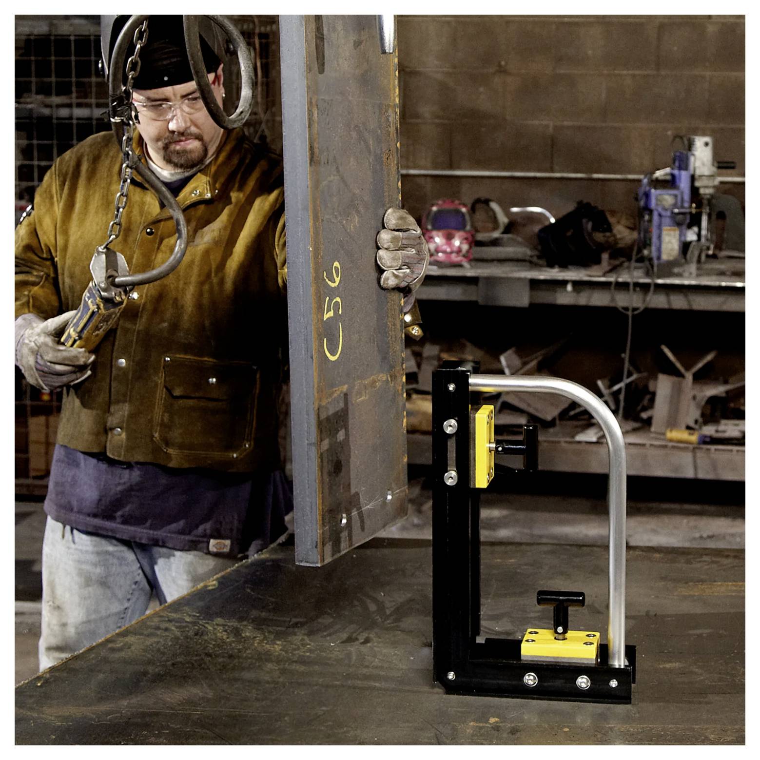 Eine Person in Schutzausrüstung verwendet einen Hebemagnet, um ein großes Metallblech in einer Werkstatt zu bewegen. Im Hintergrund sind Werkzeuge und Ausrüstung zu sehen.