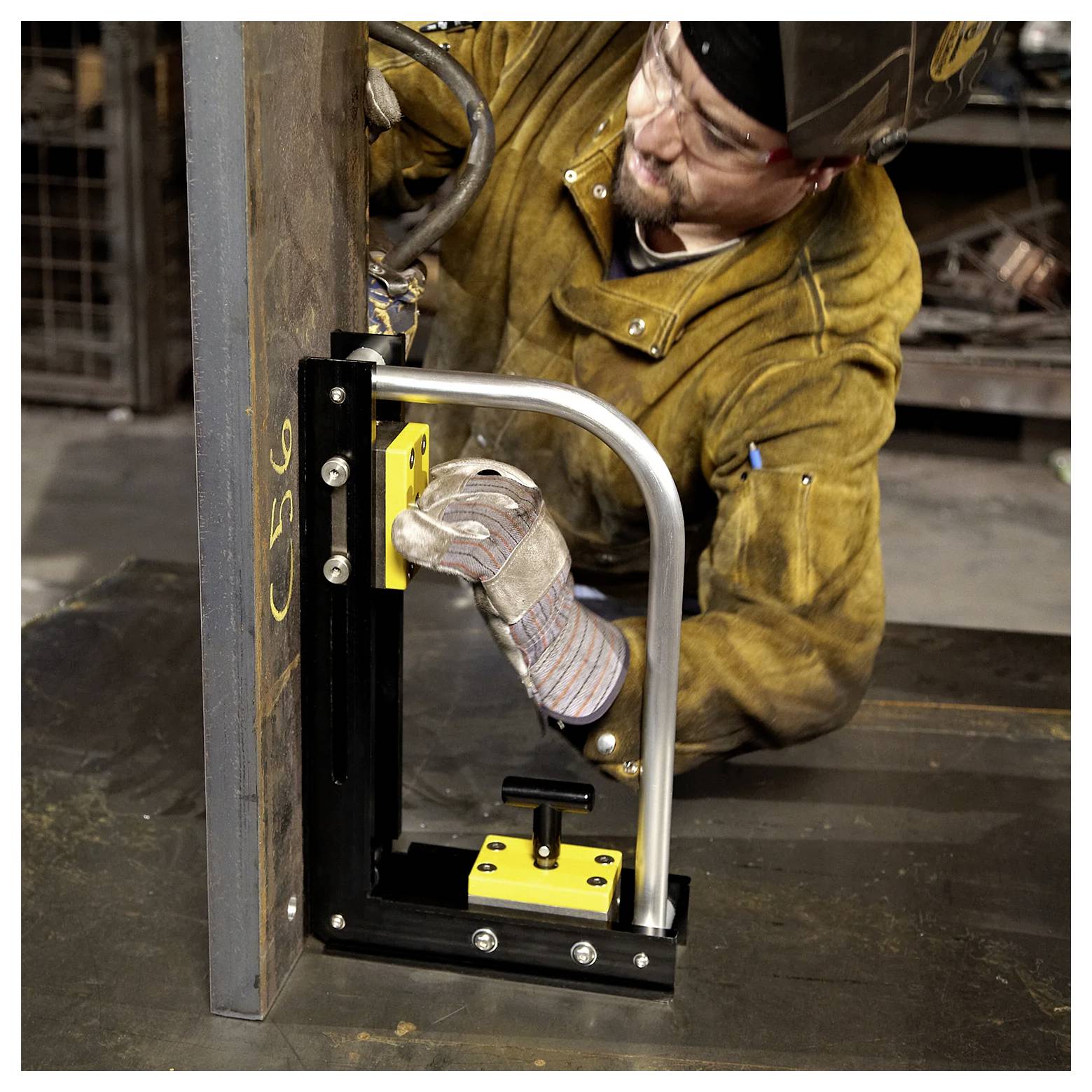 Ein Schweißer in Schutzausrüstung verwendet eine magnetische Klemme, um ein Metallblech in einer industriellen Werkstattumgebung aufrecht zu halten, während er arbeitet.