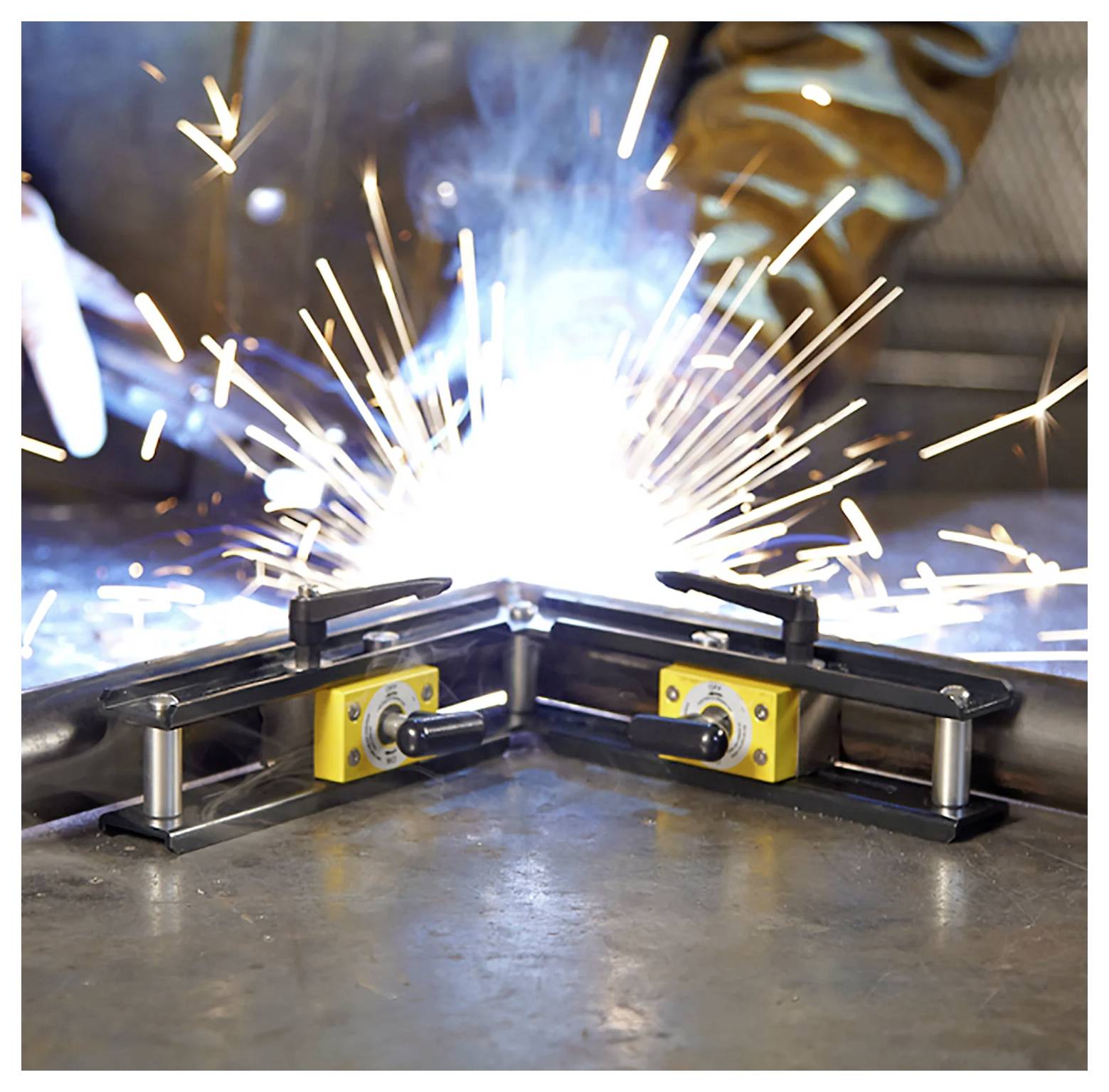 Eine Person schweißt Metallstücke auf einem Werkstatttisch, wobei helle Funken umherfliegen. Zwei magnetische Klemmvorrichtungen halten das Metall sicher an seinem Platz.