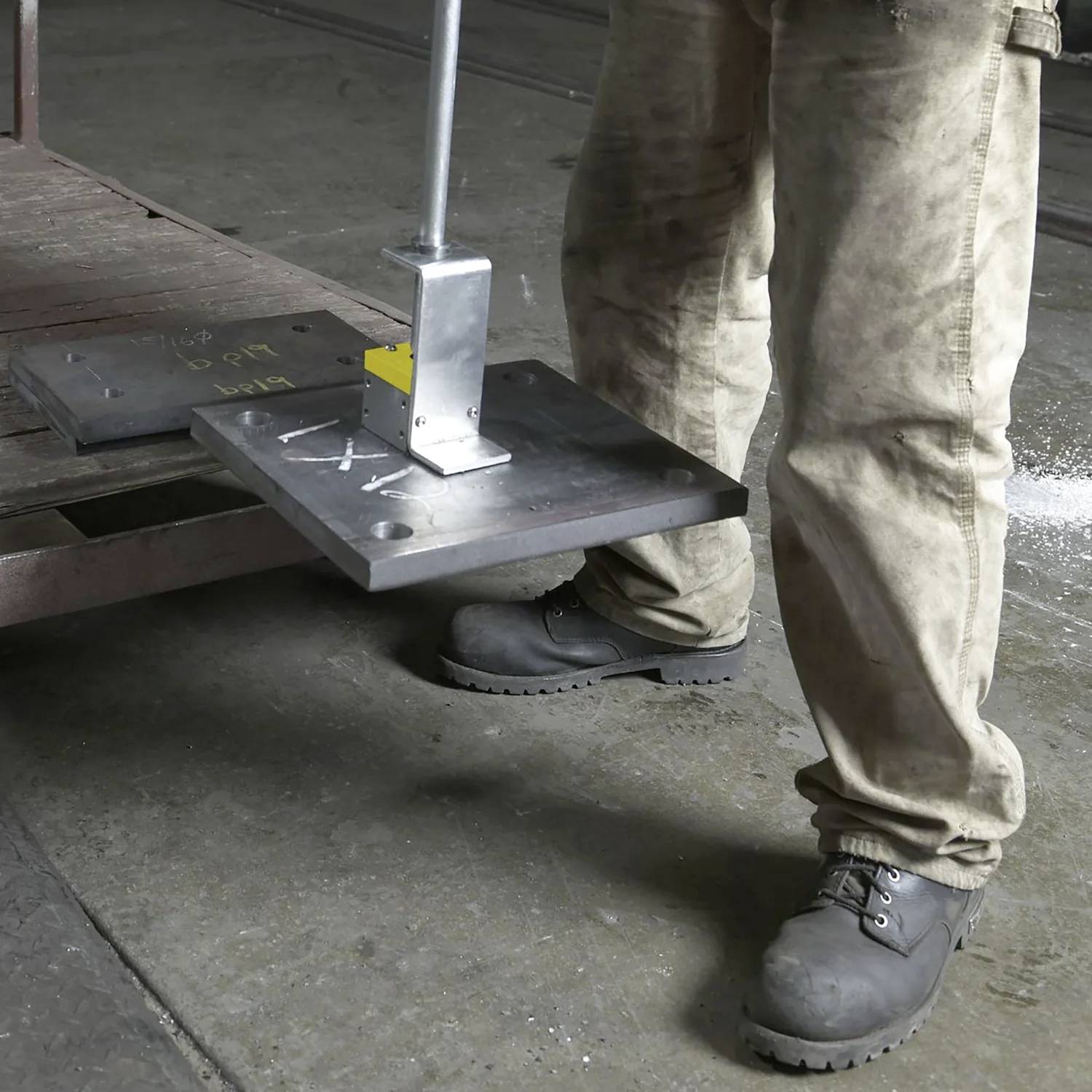 Eine Person trägt Sicherheitsschuhe und Arbeitskleidung und hebt ein Metallblech mit einem manuellen Hebewerkzeug in einer industriellen Umgebung.