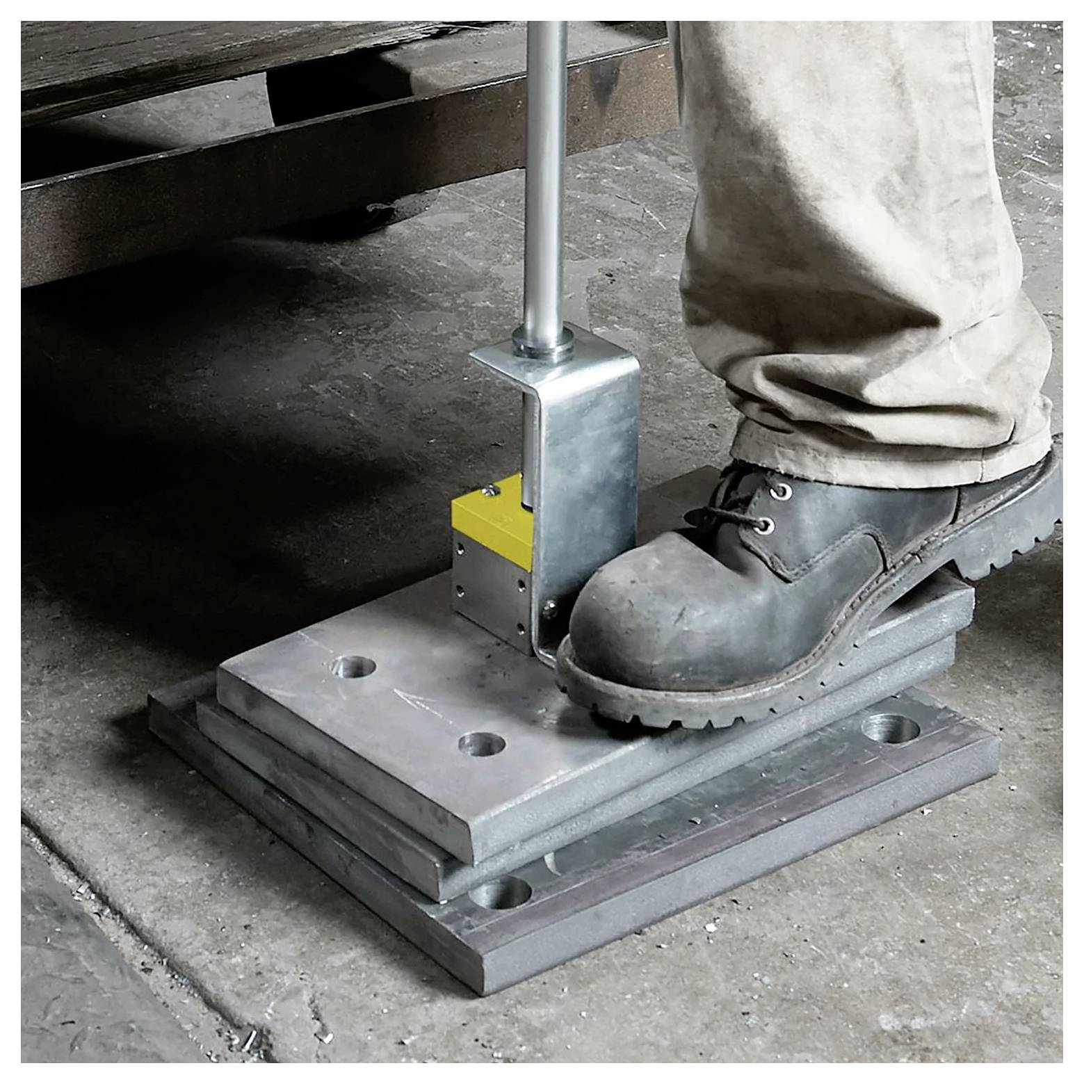 Arbeiter bedient eine Metallpresse mit dem Fuß, trägt Schutzstiefel und steht auf einem Betonboden in einem industriellen Umfeld.