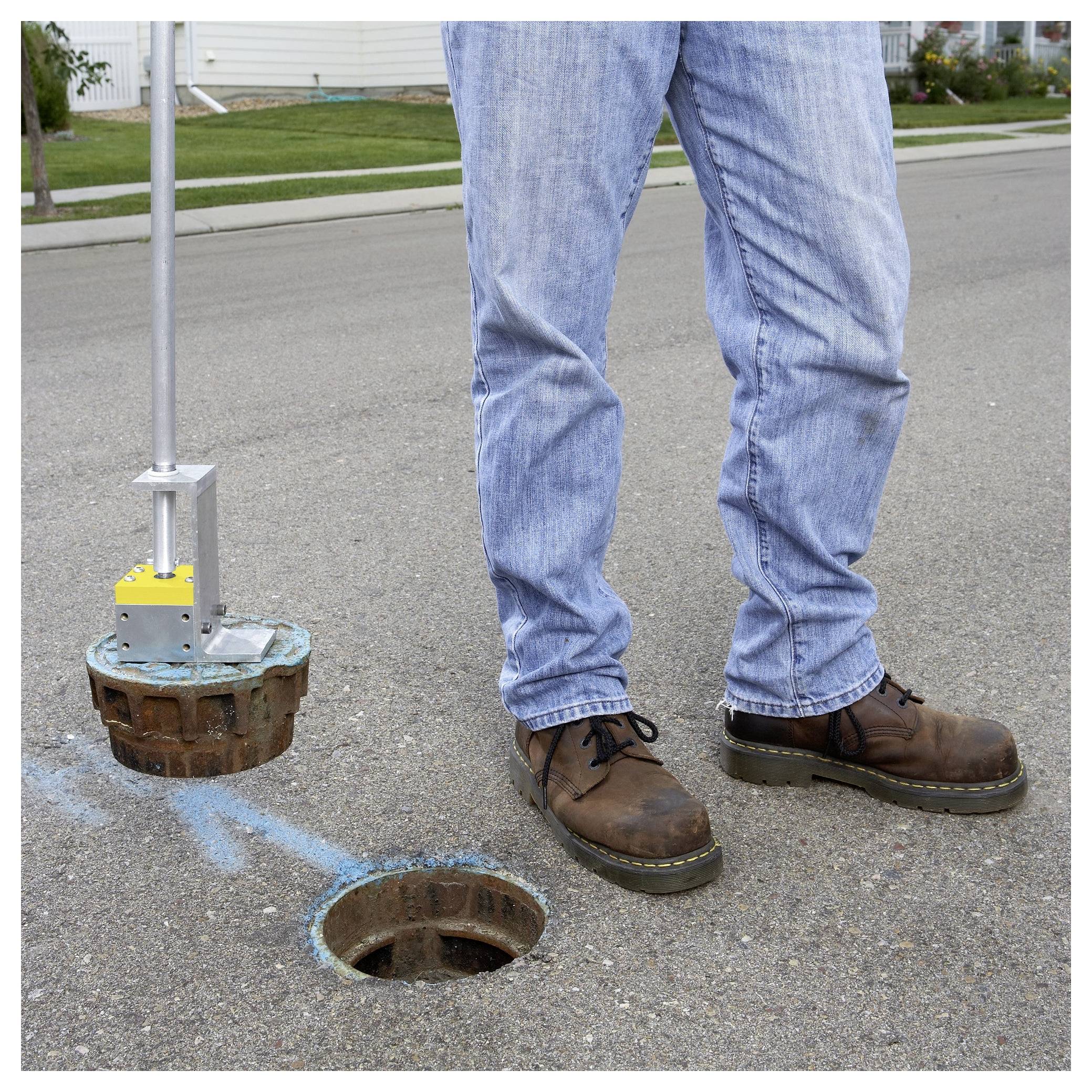 Eine Person steht neben einem offenen Kanalschacht und hält ein Werkzeug, das speziell zum Anheben des Deckels entwickelt wurde, auf einer Wohnstraße.