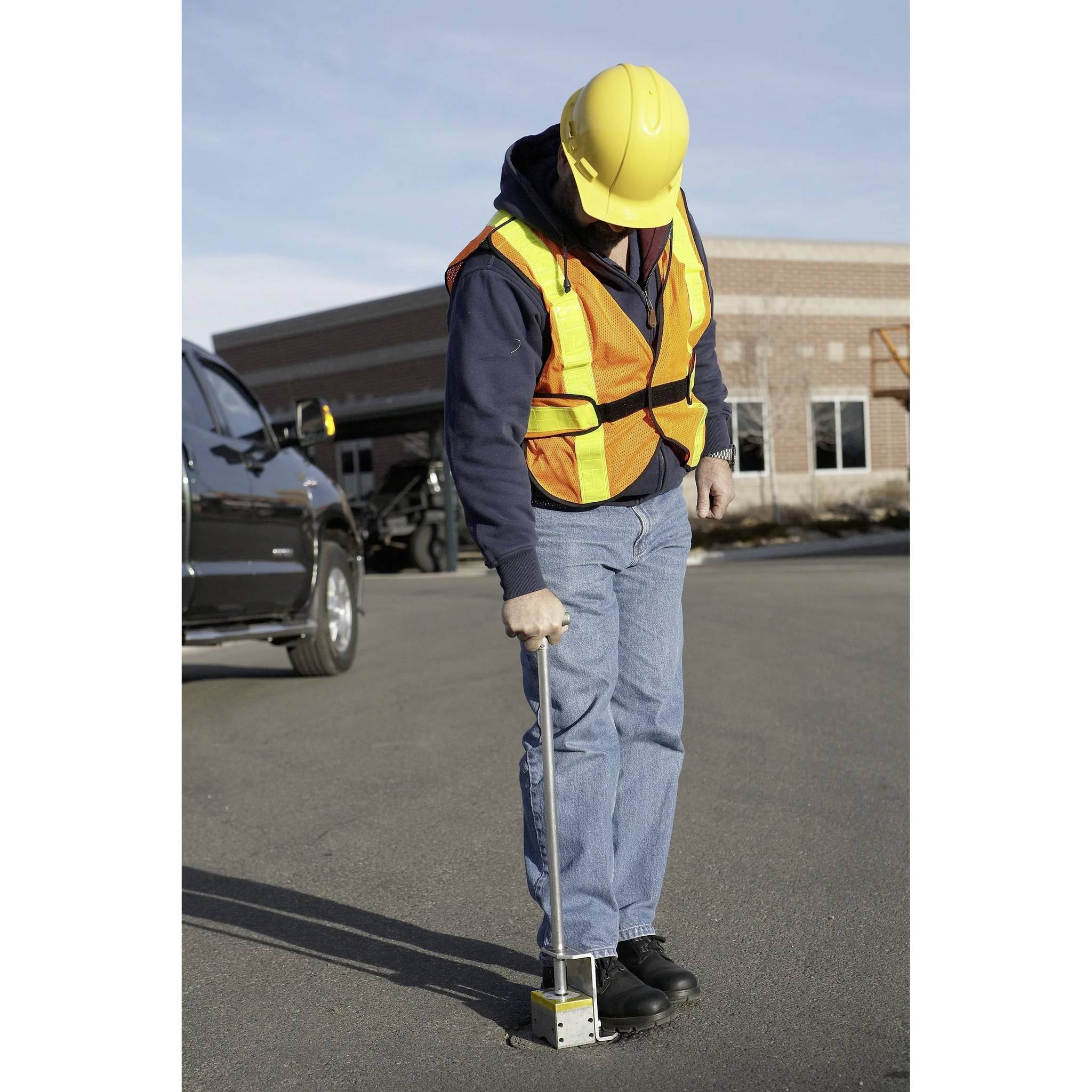 Ein Bauarbeiter mit Sicherheitsweste und Schutzhelm verwendet ein Werkzeug, um den Boden auf einer gepflasterten Oberfläche in der Nähe eines Ziegelgebäudes zu vermessen.