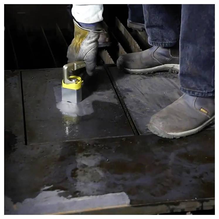 Eine Person verwendet einen schweren Magnethebemagnet, um ein Metallstück auf einer Werkstattboden zu handhaben, was auf einen Metallbearbeitungsprozess hinweist.