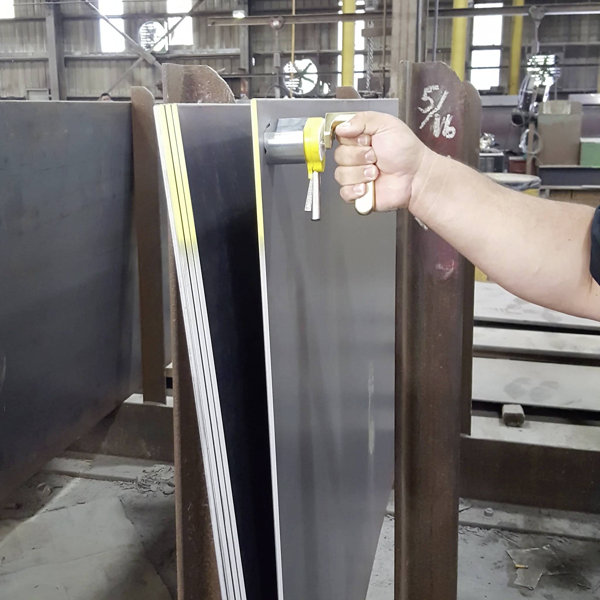 Eine Hand hält einen gelben Dickenmesser, der mehrere gestapelte Metallbleche in einer Werkstatt misst, gekennzeichnet mit '5/16'.