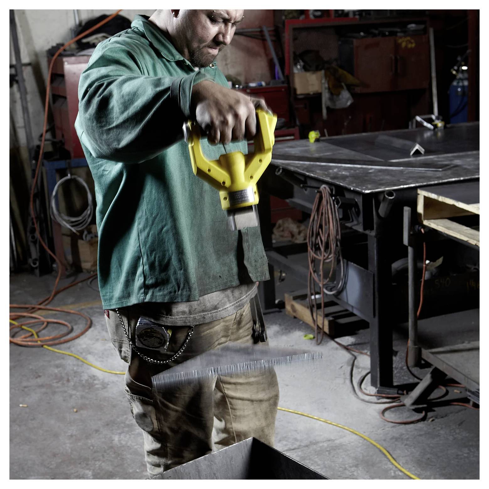Arbeiter in grünem Hemd schneidet Metallblech mit einem Elektrowerkzeug in einer Werkstatt, umgeben von Werkzeugen und Ausrüstung.