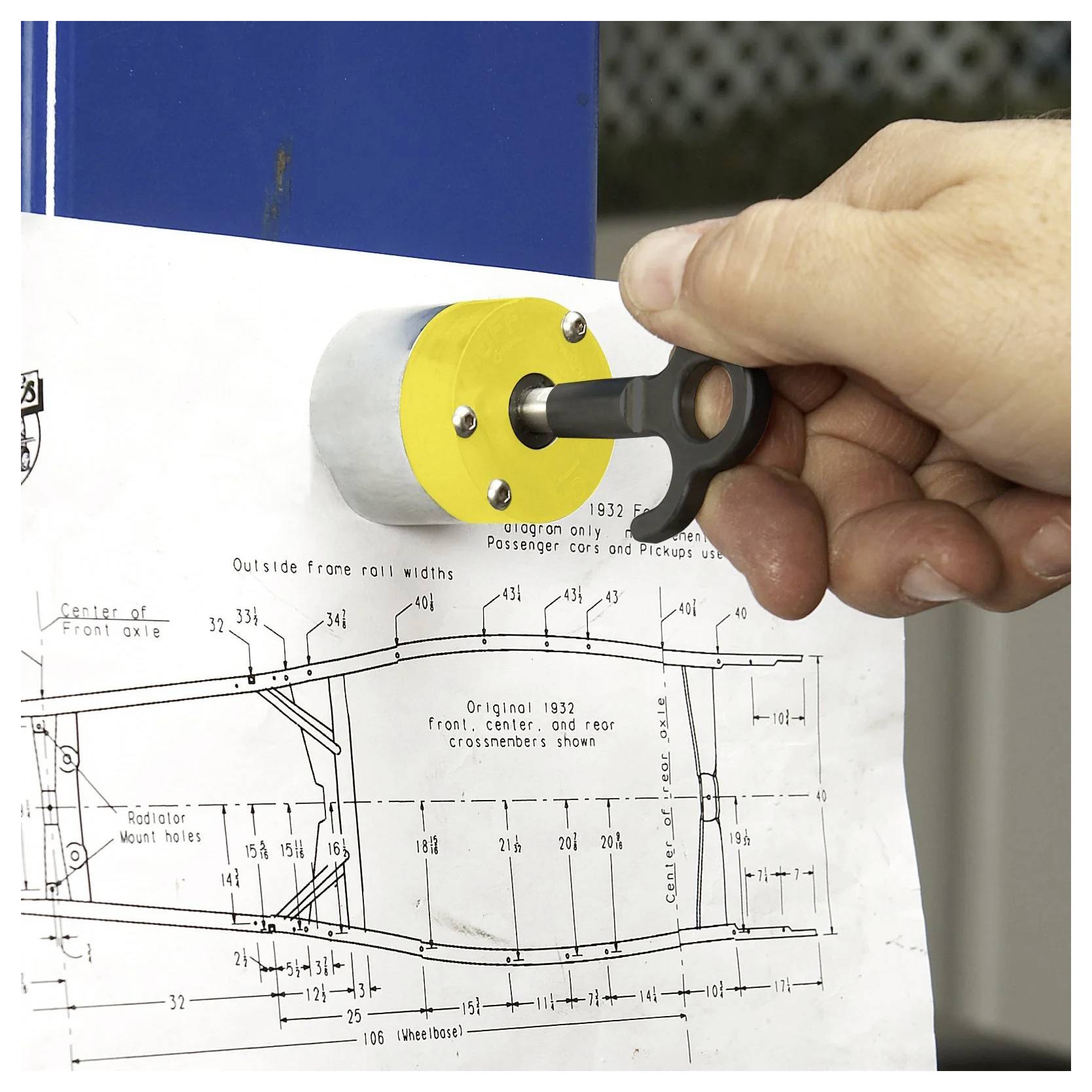 Eine Hand verwendet einen schwarzen Schlüssel mit einem gelben Halter, um eine technische Blaupause eines Fahrzeugunterbaus an eine blaue Metalloberfläche anzuheften.