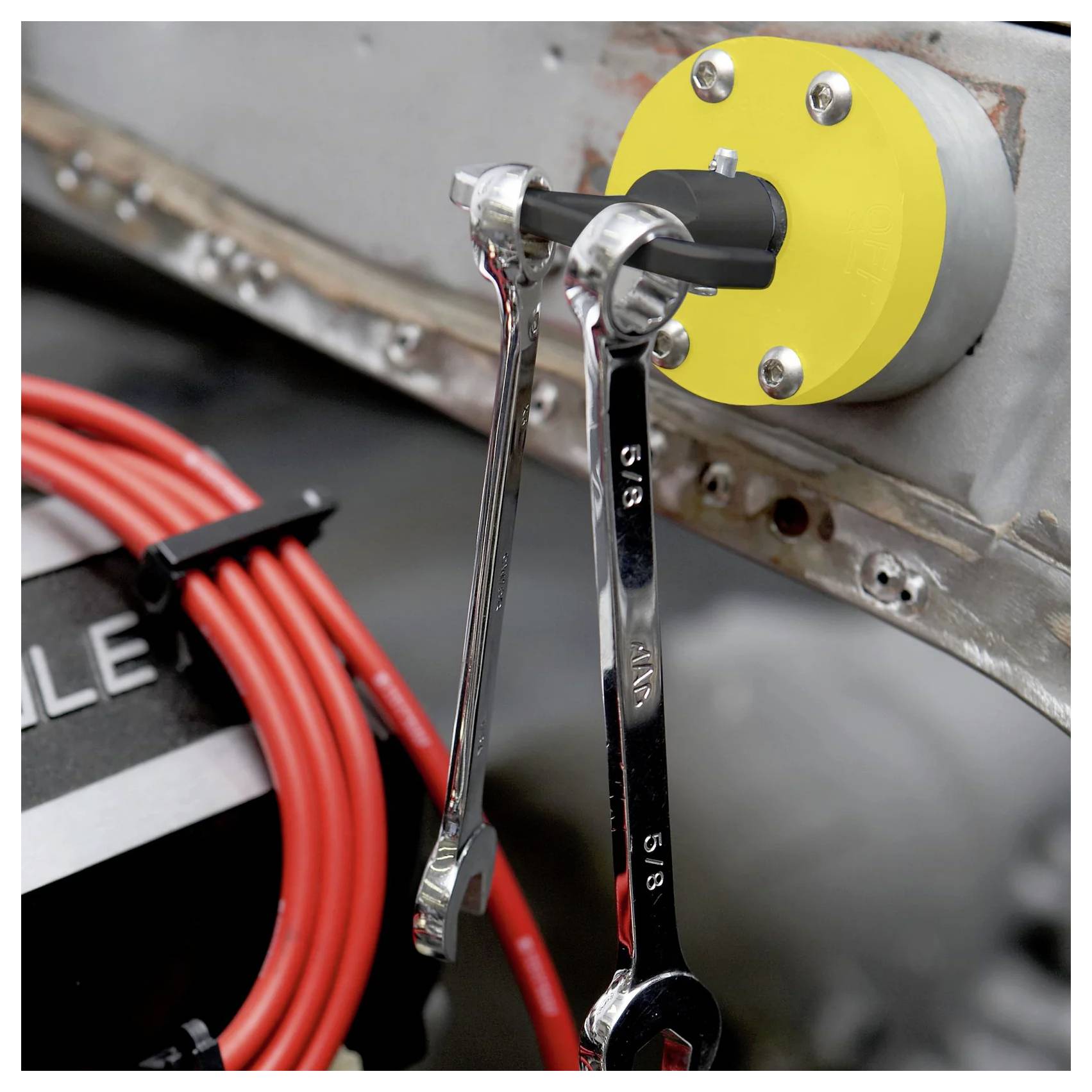 Ein gelber Magnethalter, der an einer grauen Oberfläche befestigt ist und zwei Metallschraubenschlüssel sicher hält, wobei rote Kabel im Hintergrund sichtbar sind.