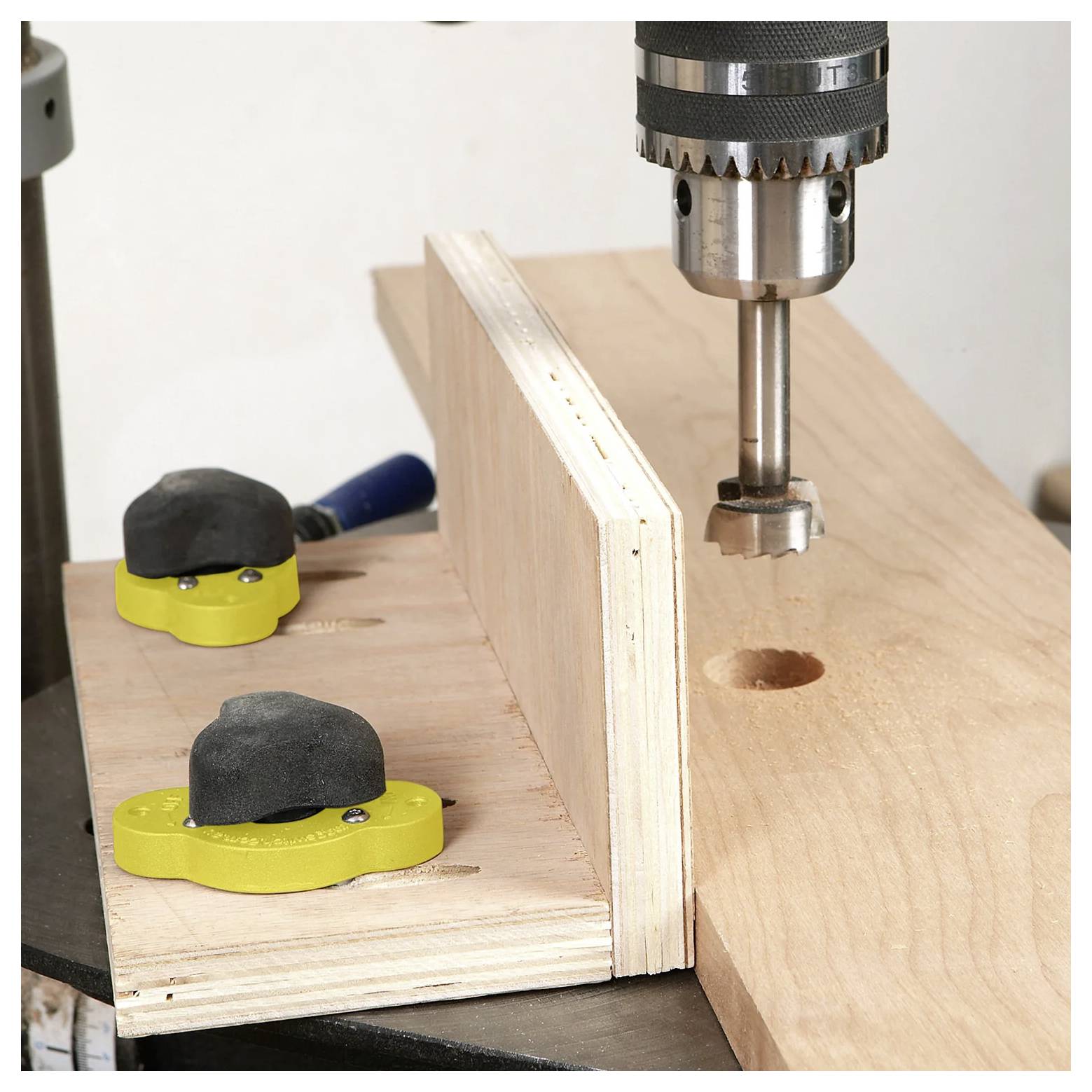 Eine Standbohrmaschine, die ein Loch in ein Holzbrett bohrt, das mit zwei gelben Schraubzwingen auf einer Werkbank sicher befestigt ist.