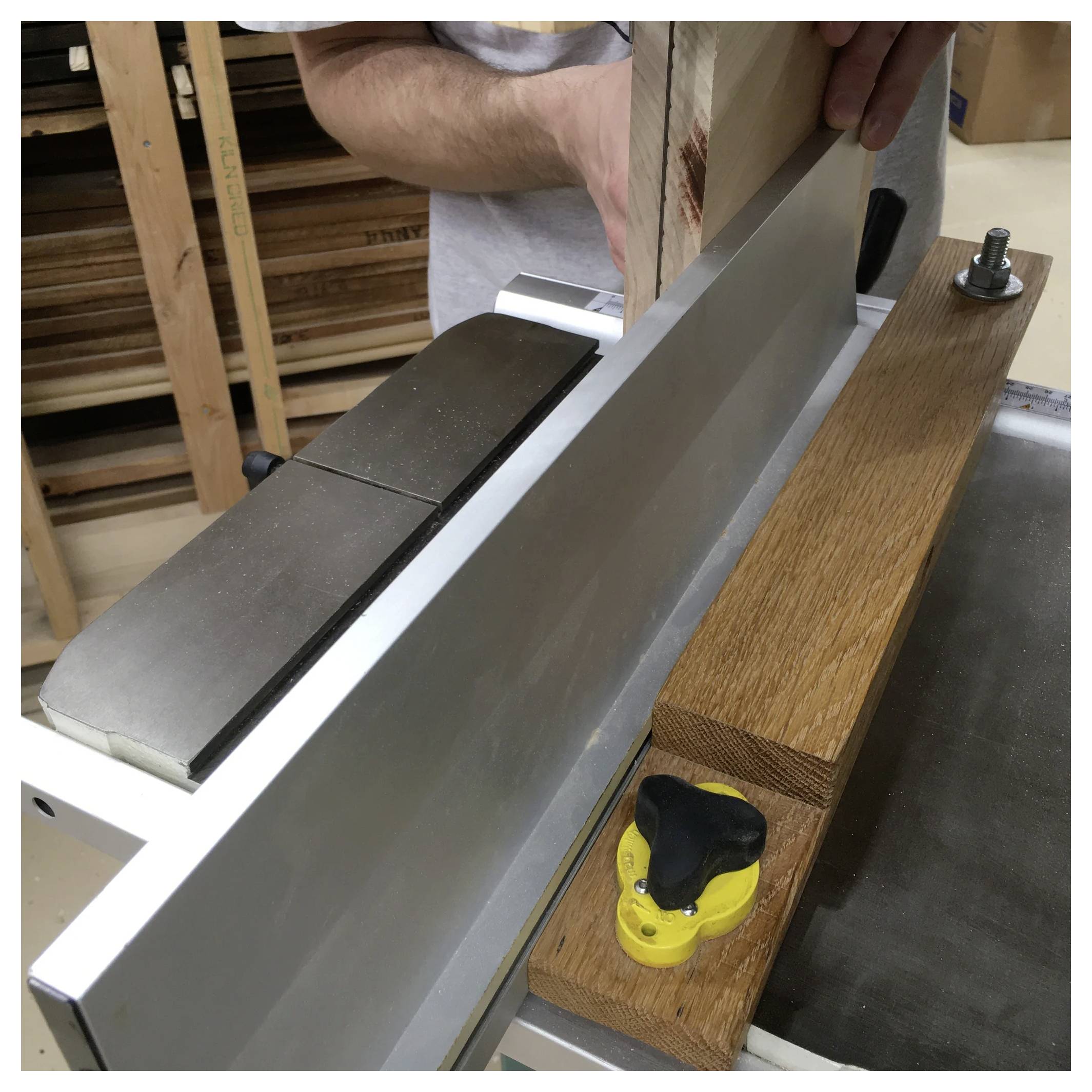 Eine Person verwendet eine Abrichthobelmaschine, um ein Holzbrett zu glätten und zu begradigen. Die Anschlagkante und das Schneidmesser der Maschine sind sichtbar, mit Holzspänen in der Umgebung.