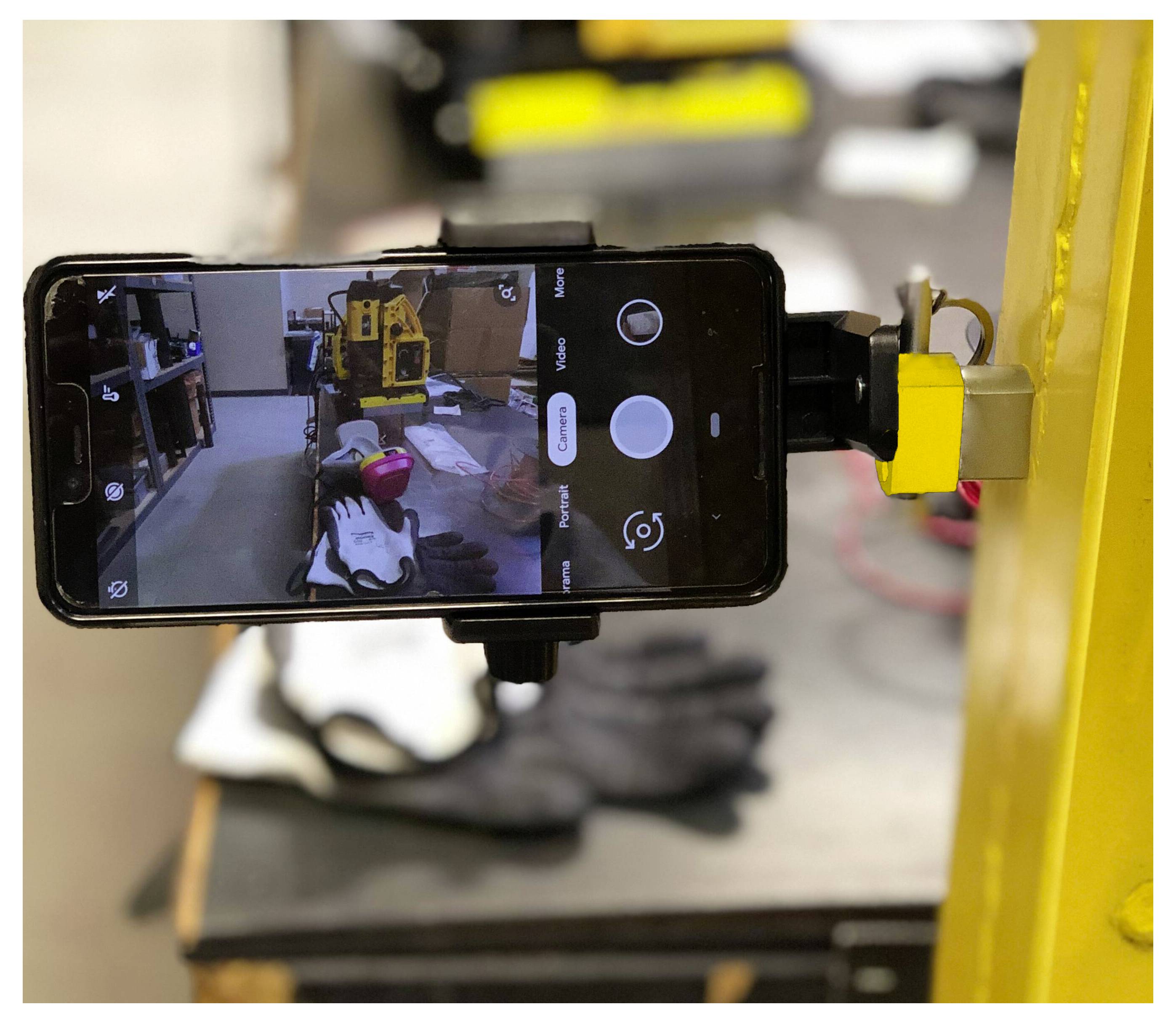 Ein Smartphone, das an gelber Maschinerieausrüstung befestigt ist, zeigt eine Kameraansicht einer Werkstatt mit Werkzeugen und einem Handschuh, der im Hintergrund auf einem Tisch liegt.