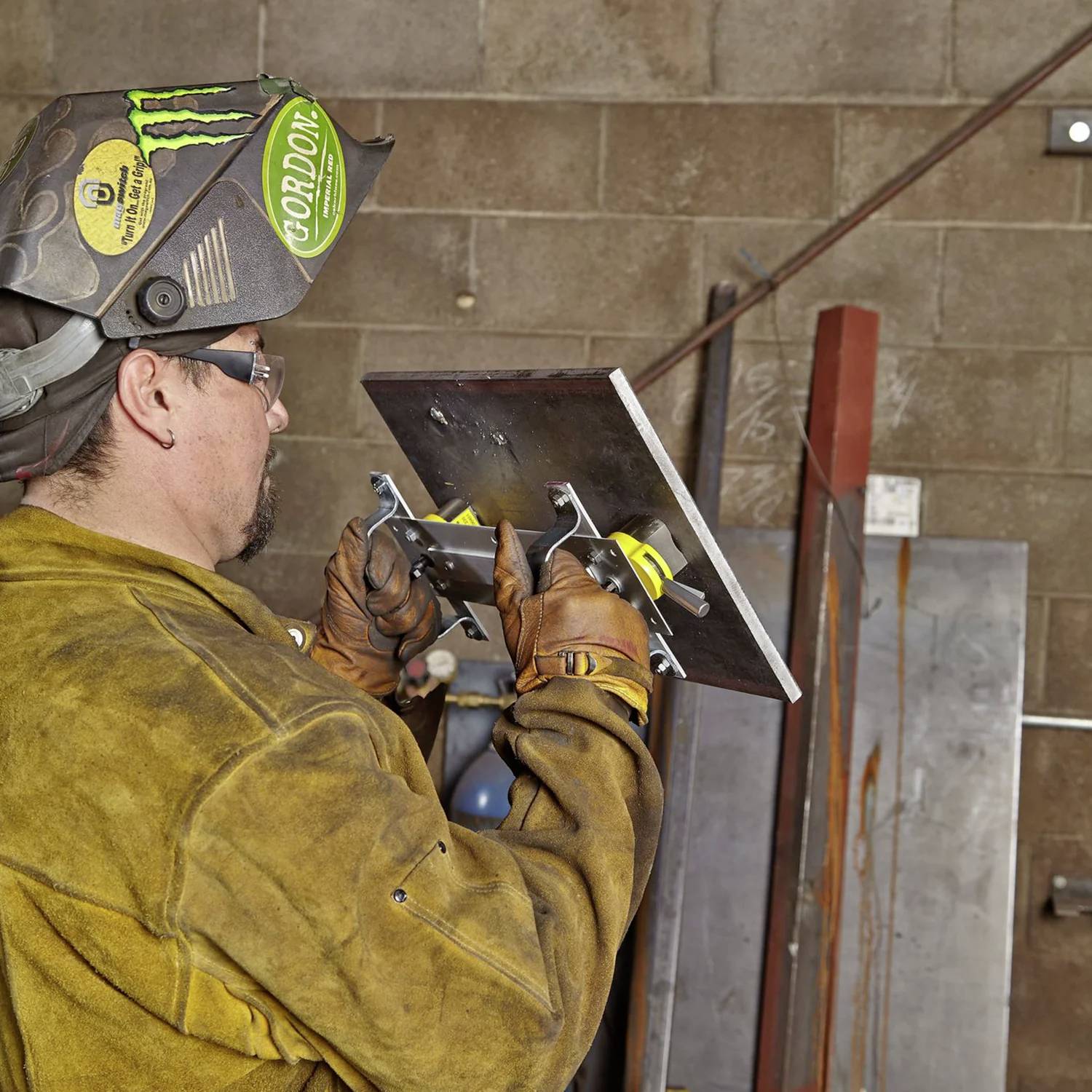 Ein Schweißer in Schutzausrüstung hält ein Metallstück mit Klammern, bereitet sich auf die Arbeit vor, vor einem Hintergrund einer Werkstattwand mit Werkzeugen.