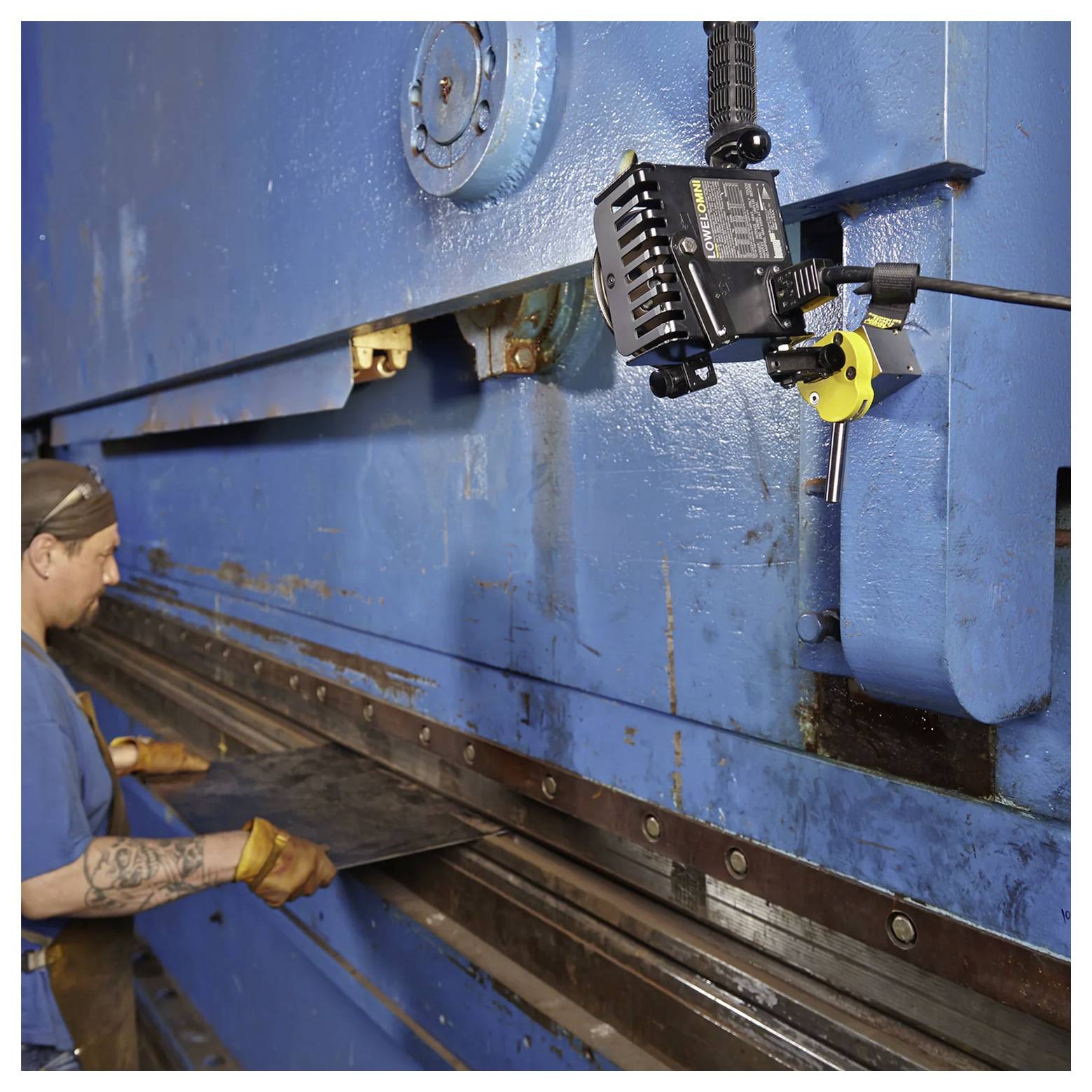 „Ein Fabrikarbeiter bedient eine große blaue Blechbiegemaschine und führt ein Metallblech in Position, wobei Steuerungsausrüstung sichtbar ist.