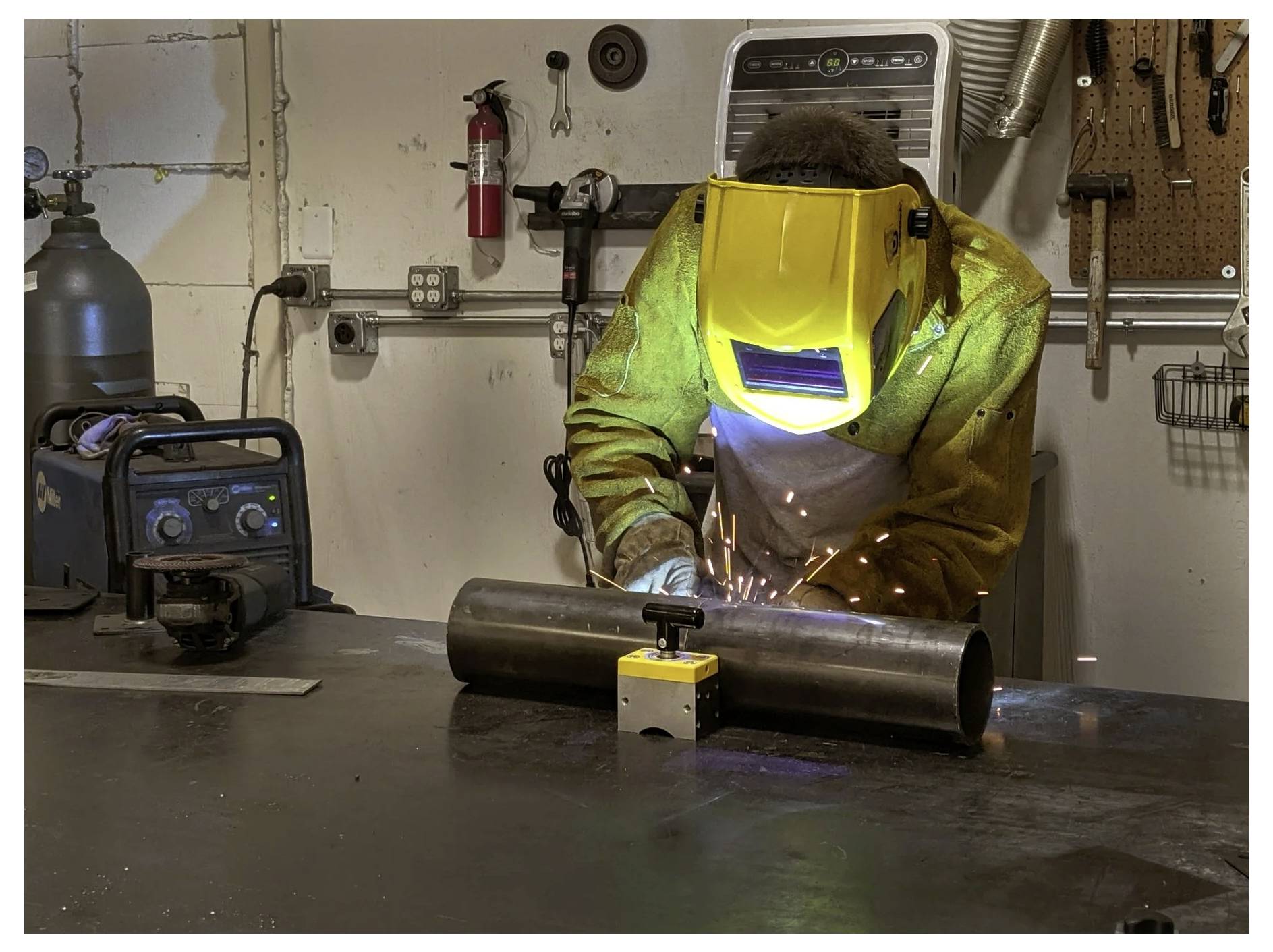 Ein Schweißer mit Schutzausrüstung und einem gelben Helm arbeitet an einem Metallrohr in einer Werkstatt, wobei Schweißfunken sichtbar sind.