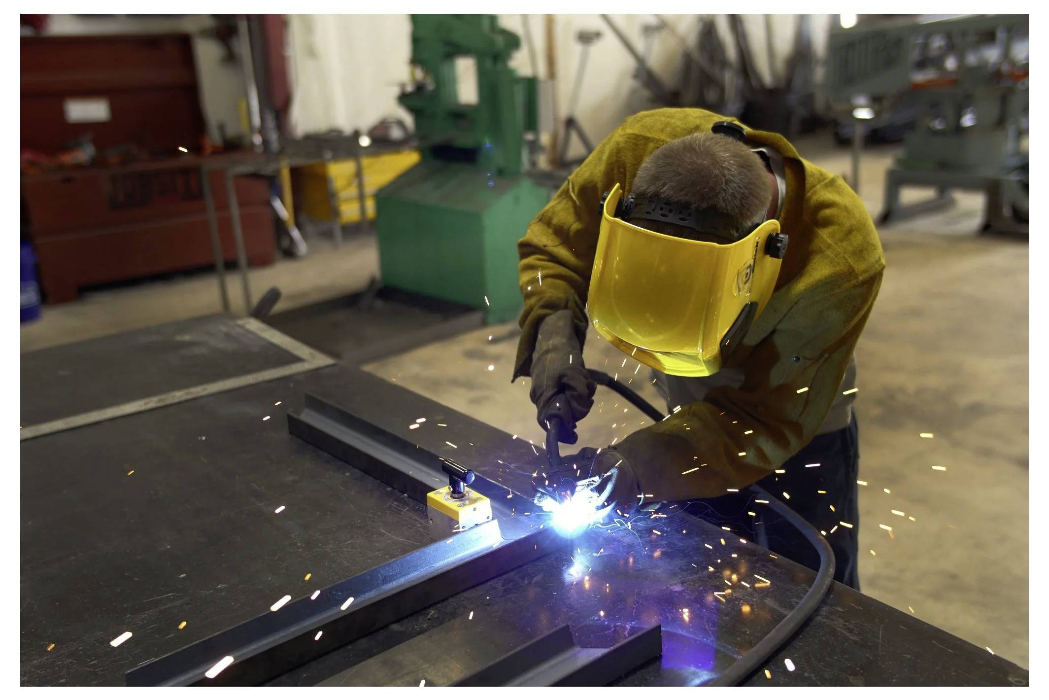 Eine Person, die Metall in einer Werkstatt schweißt und Schutzausrüstung einschließlich eines gelben Gesichtsschildes trägt; Funken sprühen während der Arbeit.