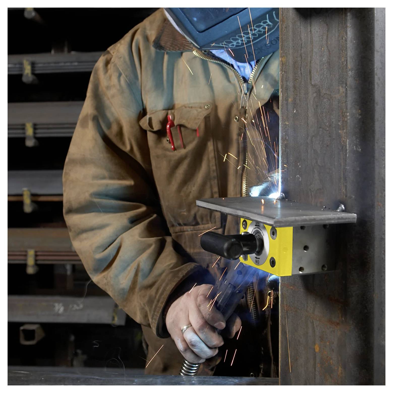 Ein Schweißer in Schutzausrüstung arbeitet an einem Stahlträger und erzeugt Funken. Im Hintergrund sind Regale mit Metallträgern zu sehen.